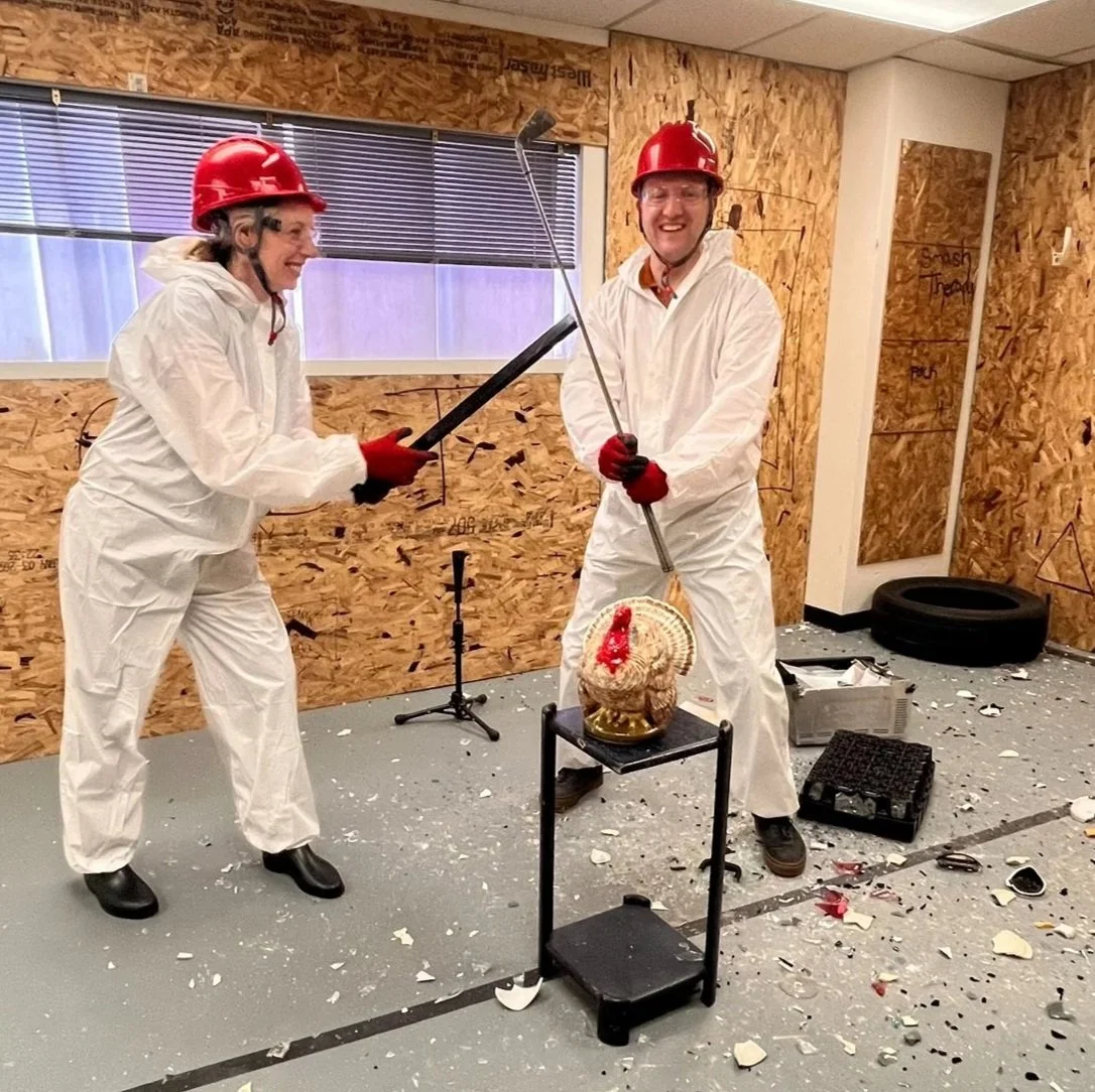 Two people in protective suits and red helmets in a rage room, smiling with weapons, surrounded by broken glass on the floor.