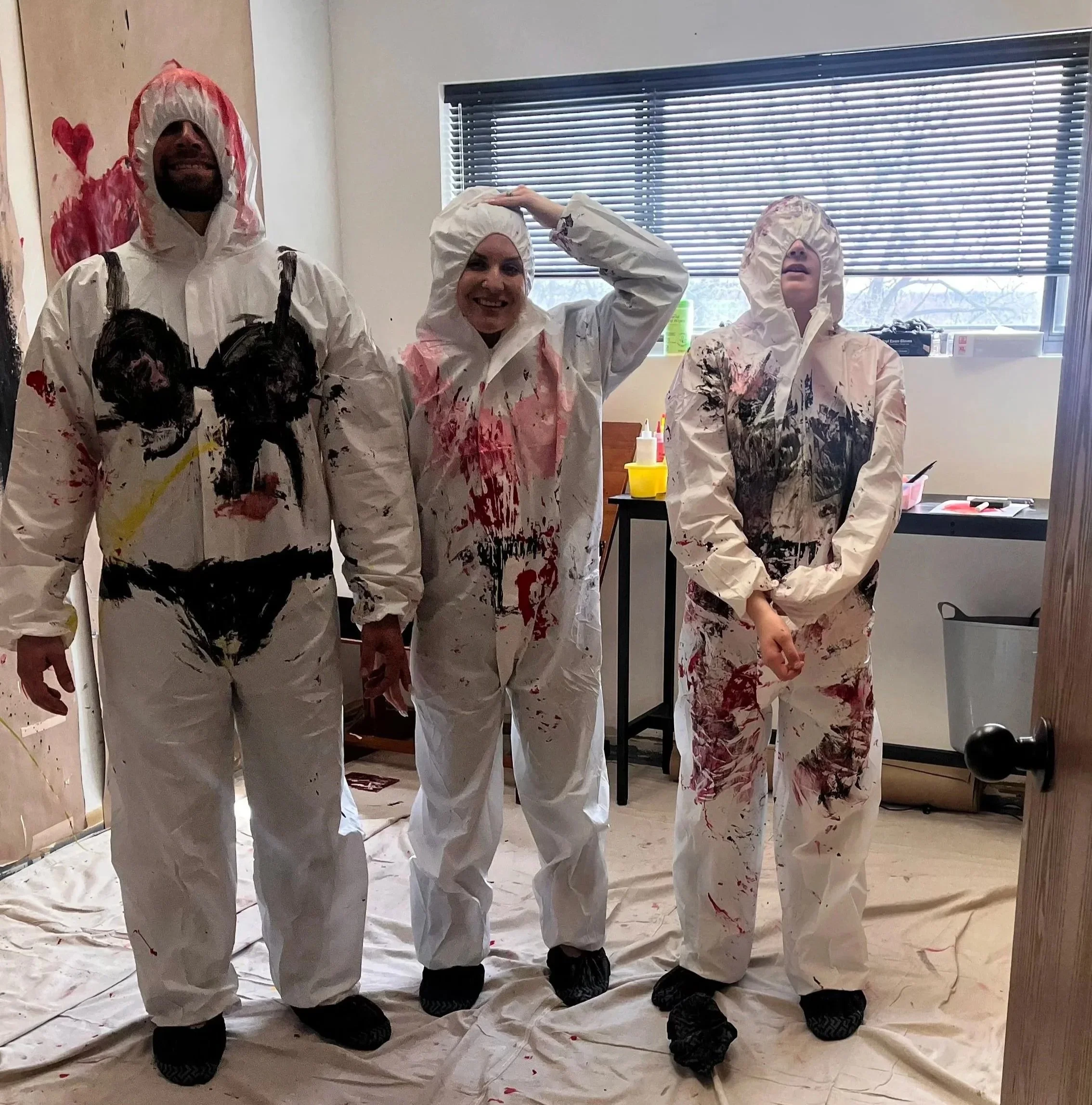 Three people wearing white paint-splattered coveralls standing on a covered floor in a room with art supplies. Paint Splatter Room