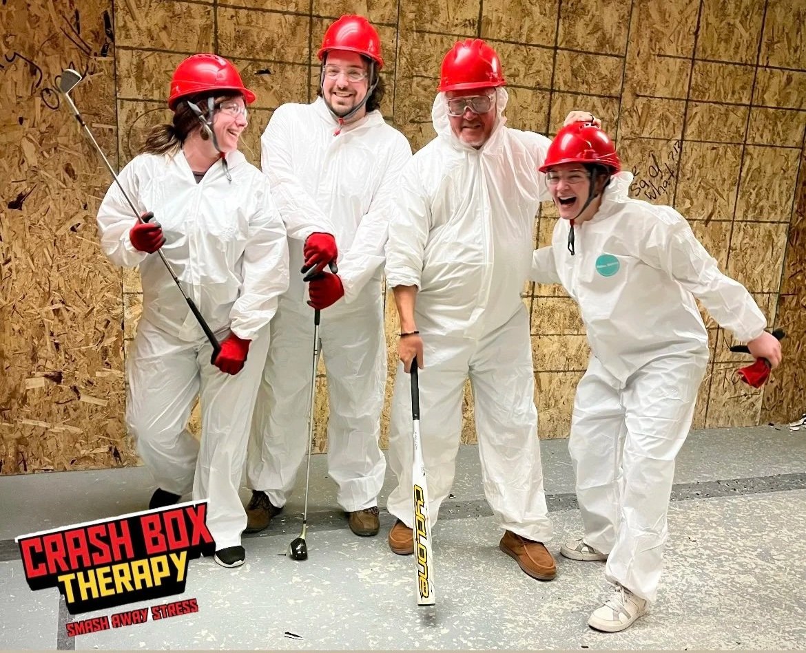 Five people in protective white coveralls and red helmets playing miniature golf inside a plywood-enclosed room, laughing and smiling. One woman is holding a golf club, and the others are holding golf balls or clubs. A logo in the bottom left corner 