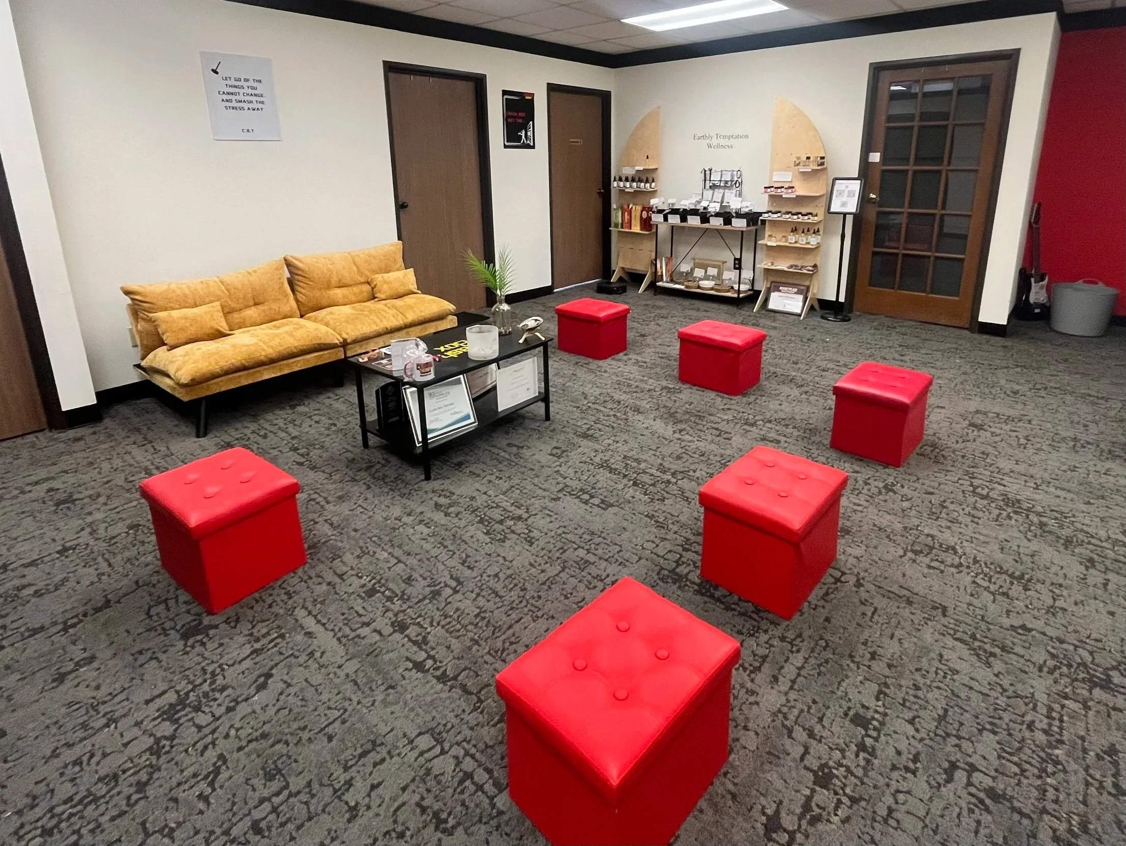 A cozy waiting area with a mustard yellow sofa, a black coffee table, and red ottomans on a patterned gray carpet. There's a display of wellness products on the wall and a wall sign that reads 'Earthly Temptation Wellness.' The room has white walls, 