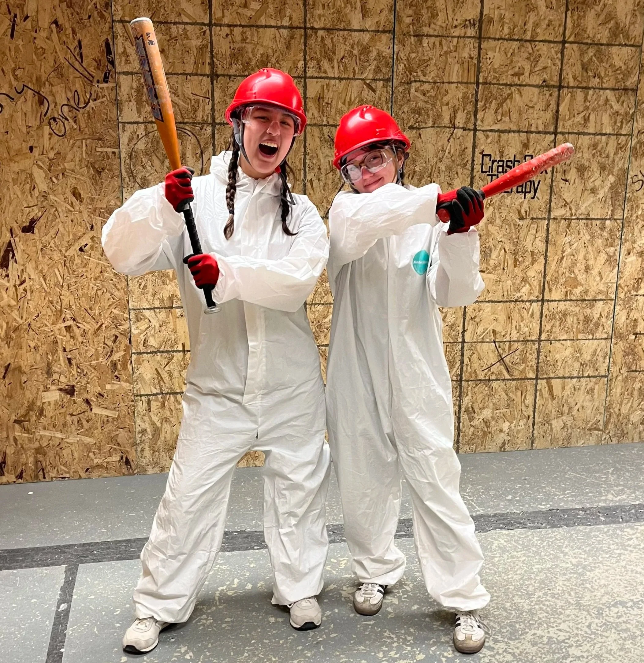 Two young women wearing white protective suits, red helmets, safety goggles, and gloves, holding baseball bats and posing inside a Crash Box Therapy Smash room with wood wall panels in the background.