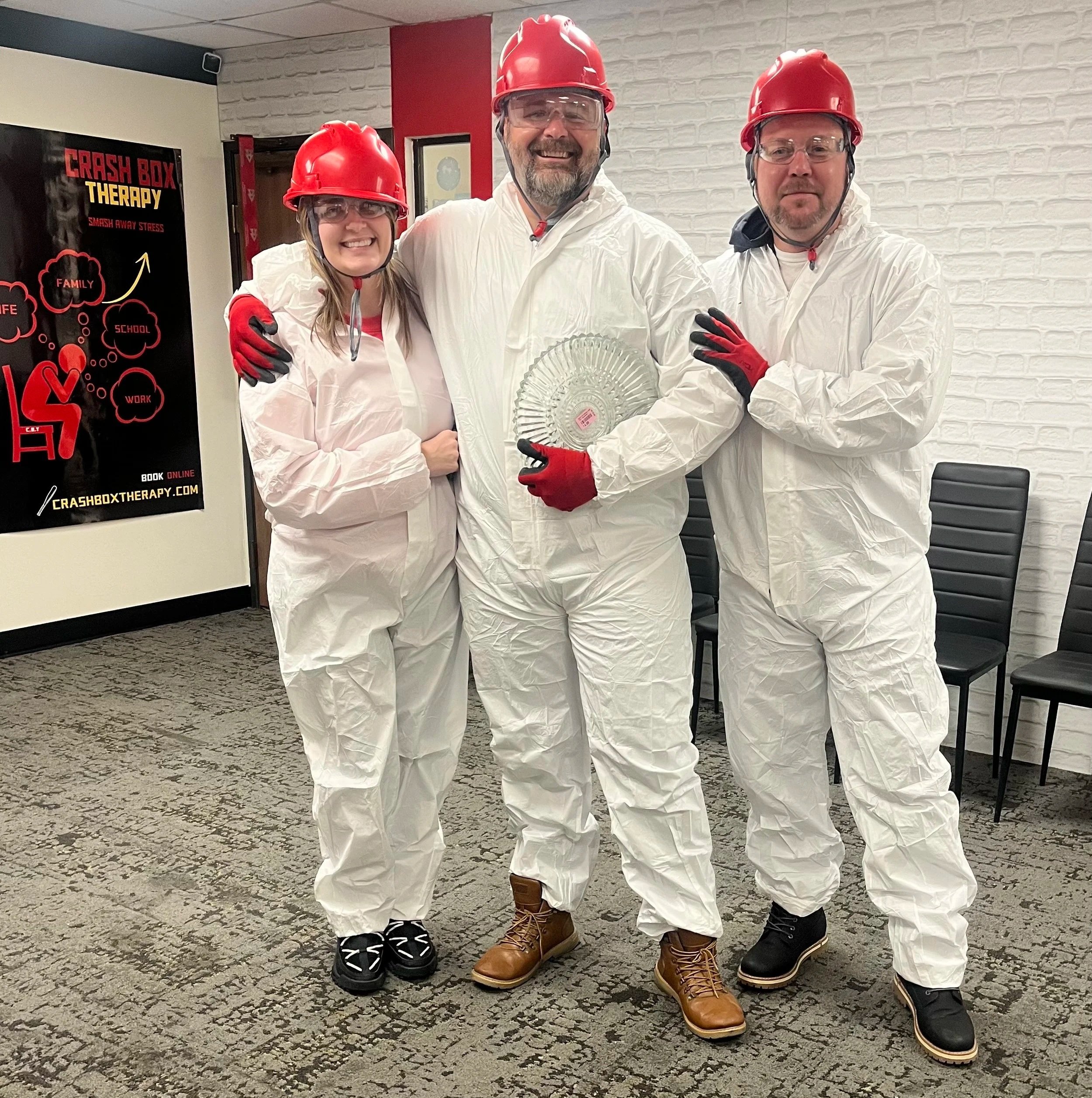 Three people wearing white protective suits, red safety helmets, and gloves standing indoors on a carpeted floor, smiling at the camera. The person in the middle is holding a glass award. There is a black poster on the wall to the left with red and y