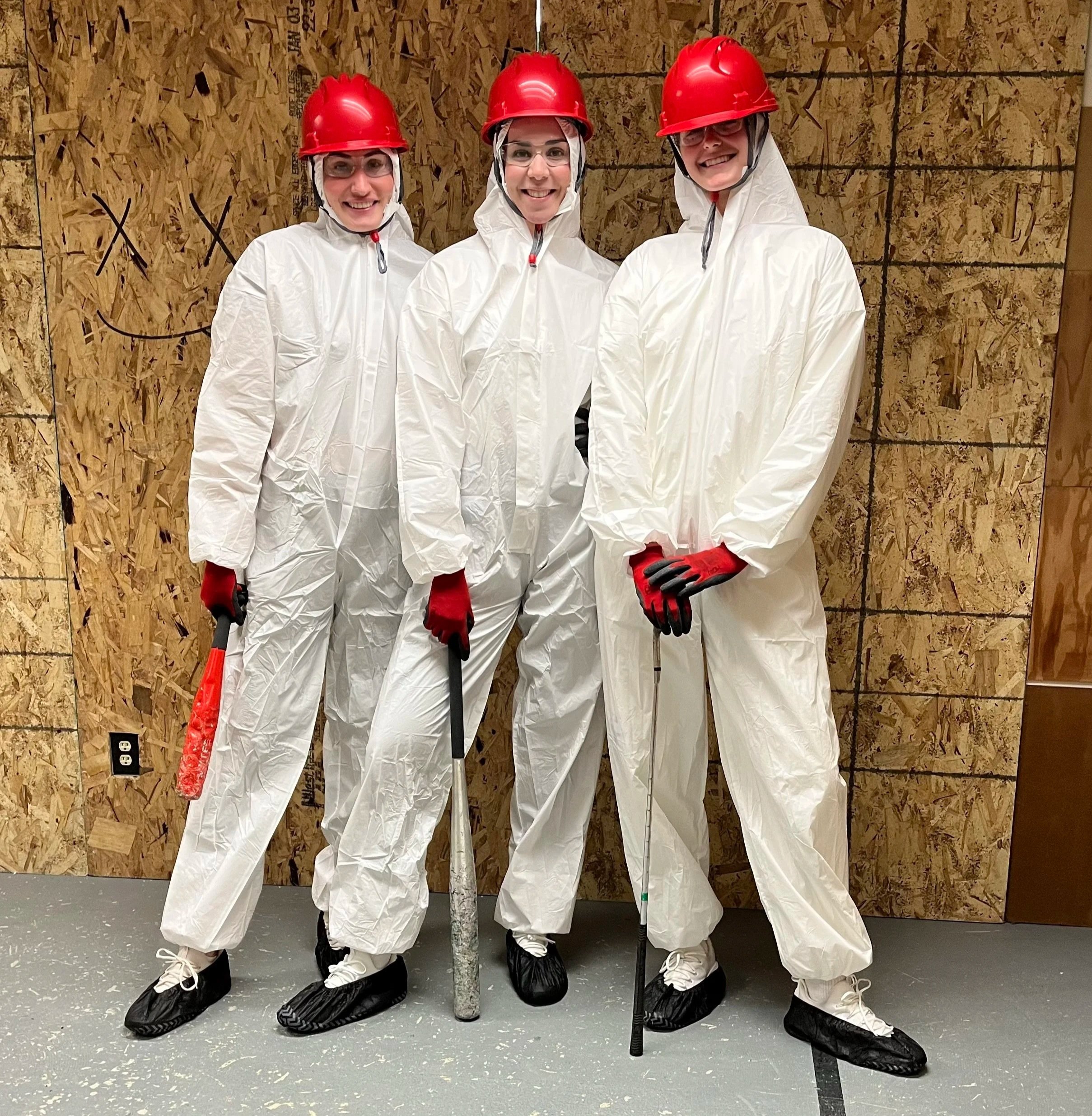 Three women wearing white protective suits, red hard hats, safety glasses, black shoe covers, and red gloves standing inside Crash Box Therapy with OSB wall sheathing and a black outline drawn on it. They are smiling and standing close together.