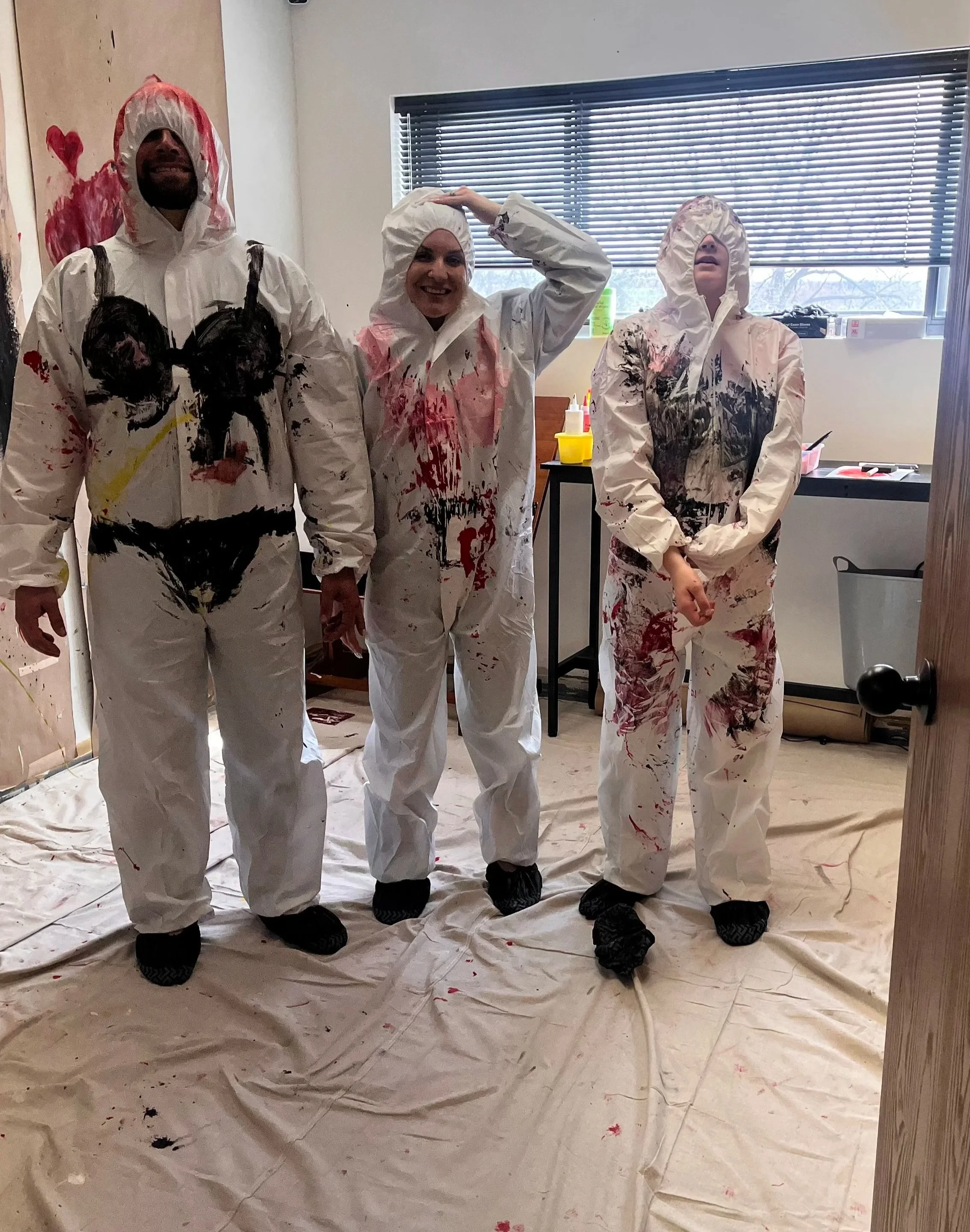 Three people dressed in white coveralls with paint splatters, standing in a room with a beige tarp on the floor and a window with blinds behind them.