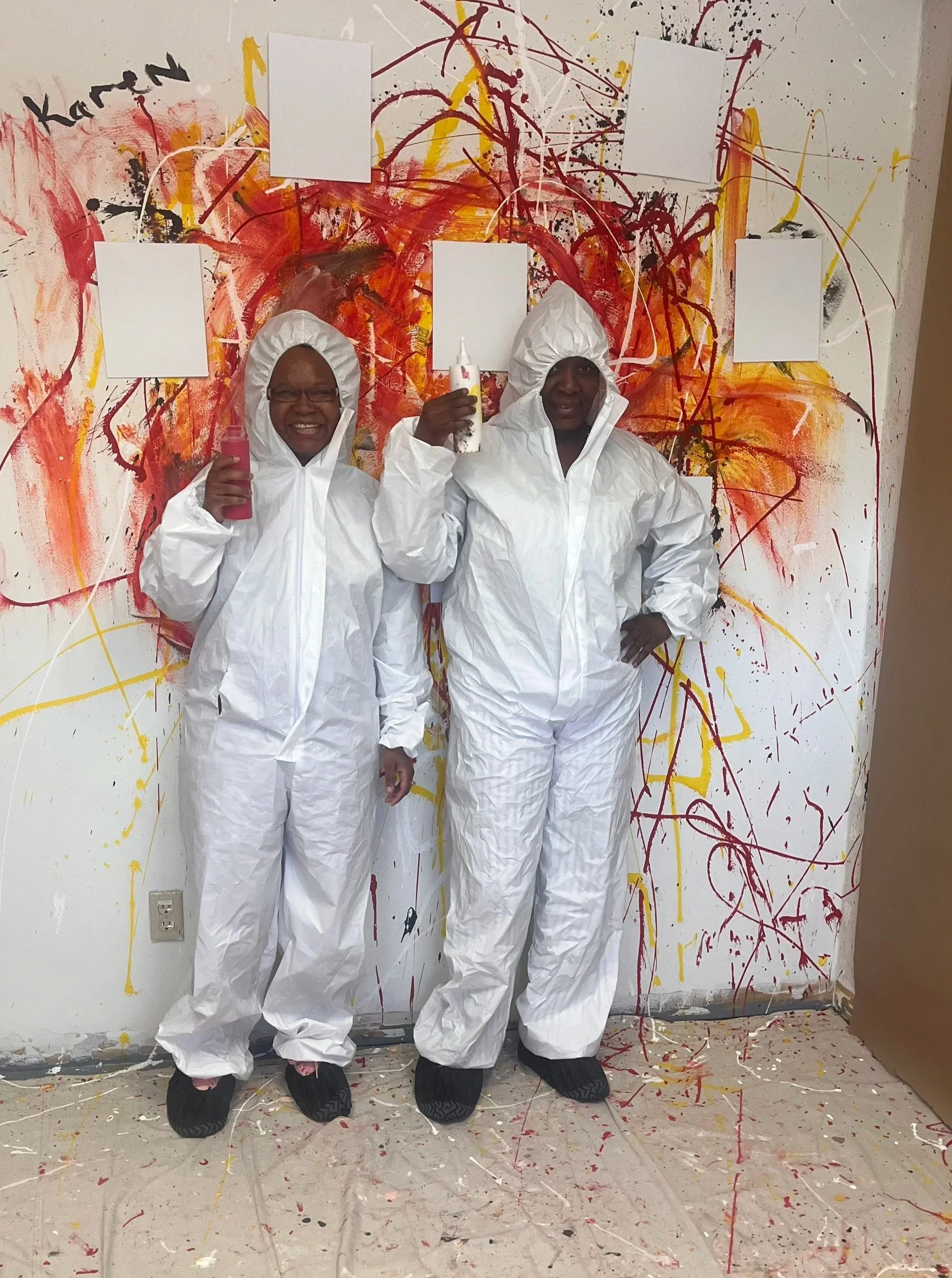 Two women dressed in white protective suits stand in front of a colorful, abstract splatter-painted wall, holding up bottles. The wall features splashes of red, yellow, black, and white paint, and the floor below has paint splatters as well.