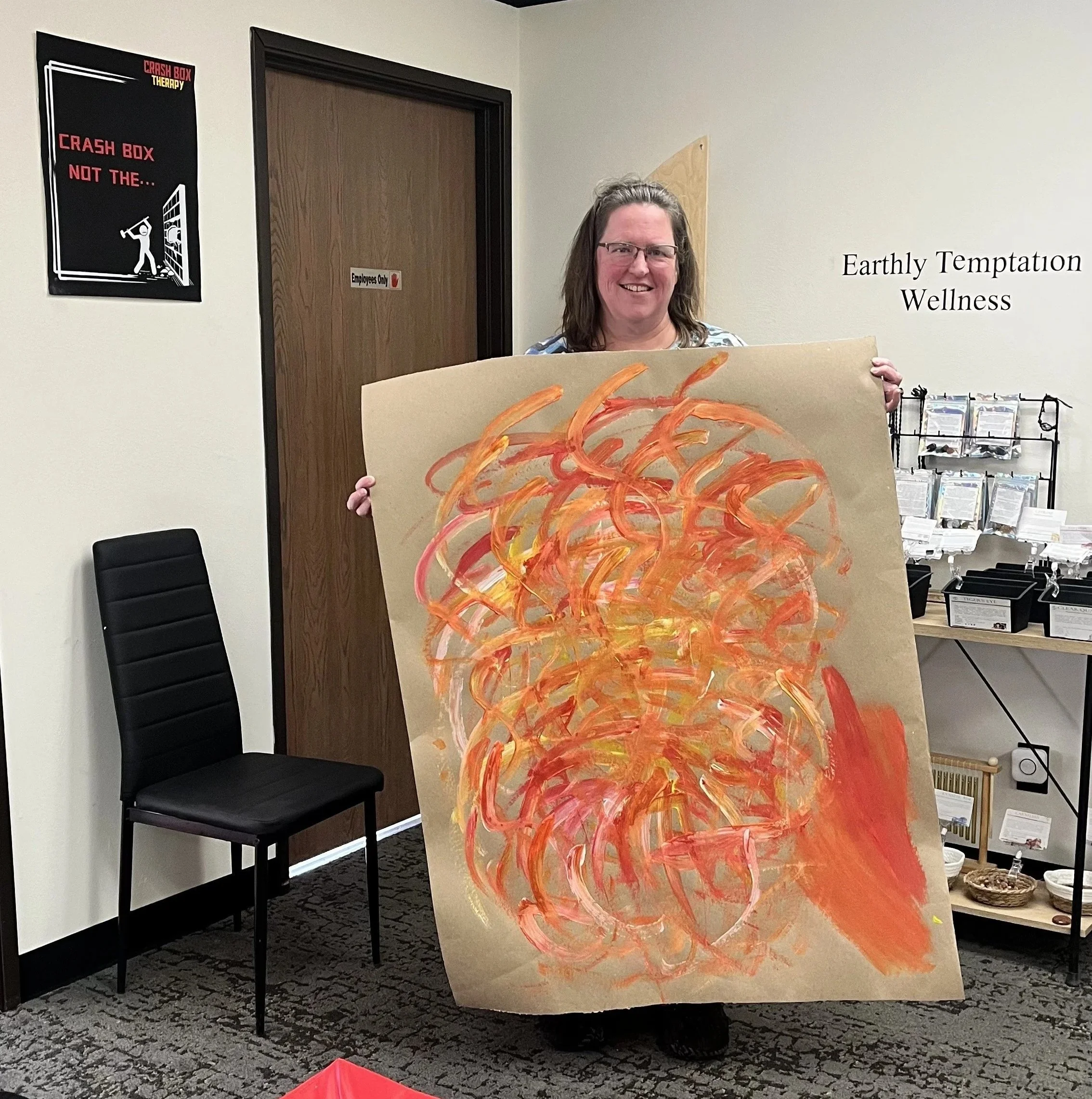 A woman with glasses and shoulder-length hair holding a canvas with an abstract orange and yellow swirl painting. The background shows a sign that reads 'Earthly Temptation Wellness' and a sign on a door that says 'Employees Only.' There is a black c