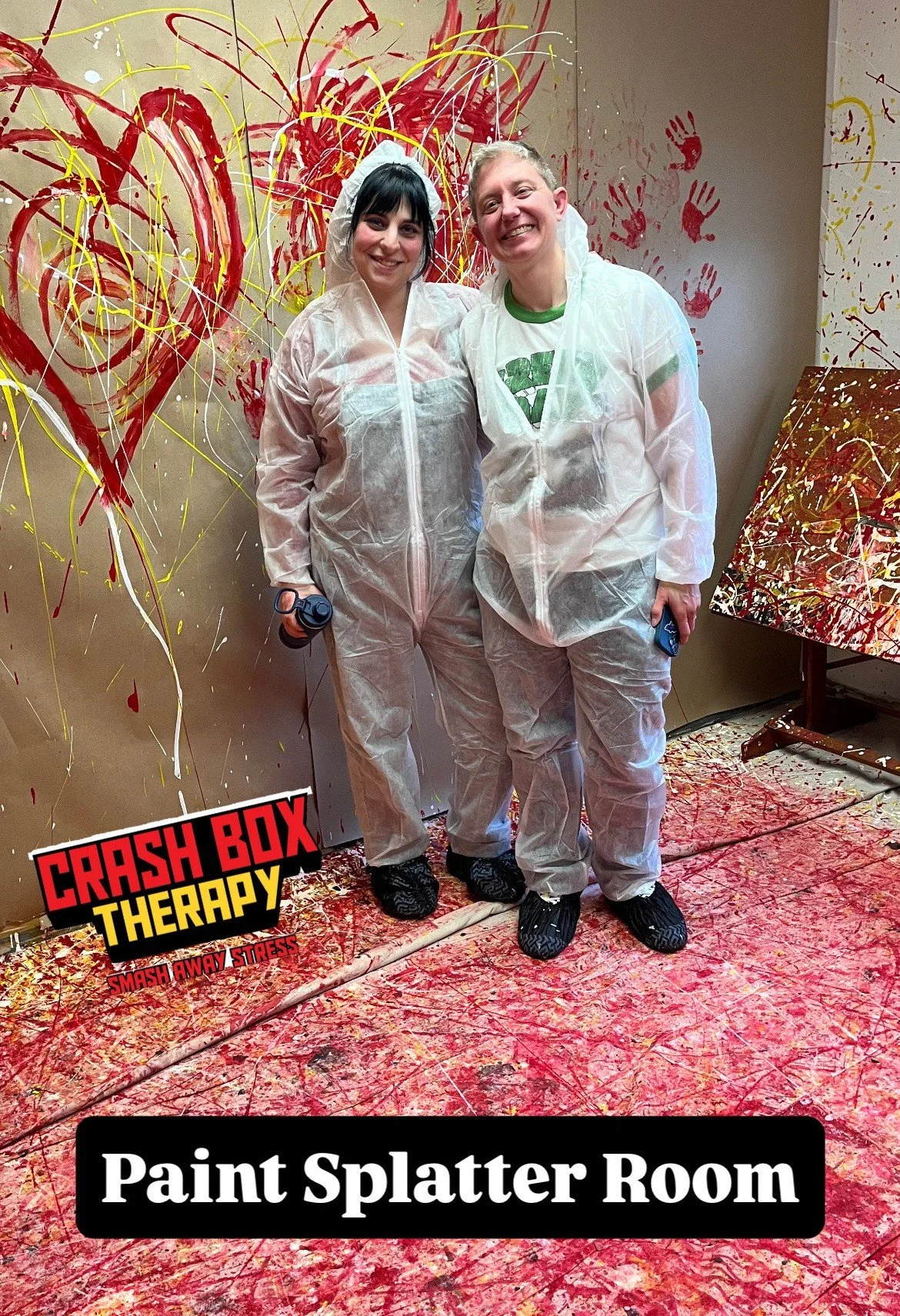 Two people in protective suits standing in a room with red, yellow, and white paint splatters on the walls and floor, in front of a large abstract heart-shaped painting. There is a sign that says 'Crash Box Therapy' and 'Paint Splatter Room'.