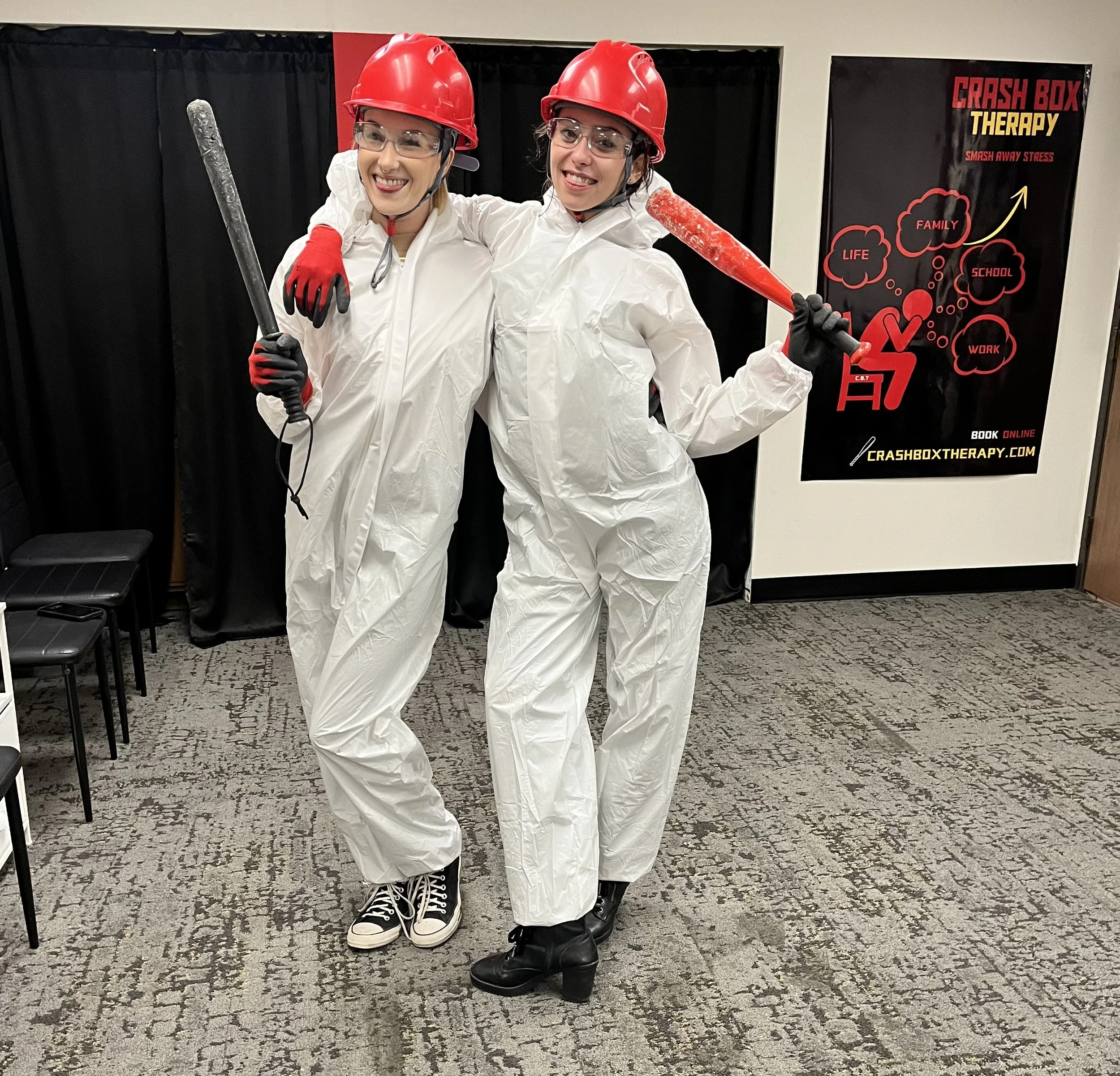 Two people wearing white protective suits, red helmets, clear safety glasses, and gloves, holding large mock baseball bats, smiling at the camera during a crash box therapy event.