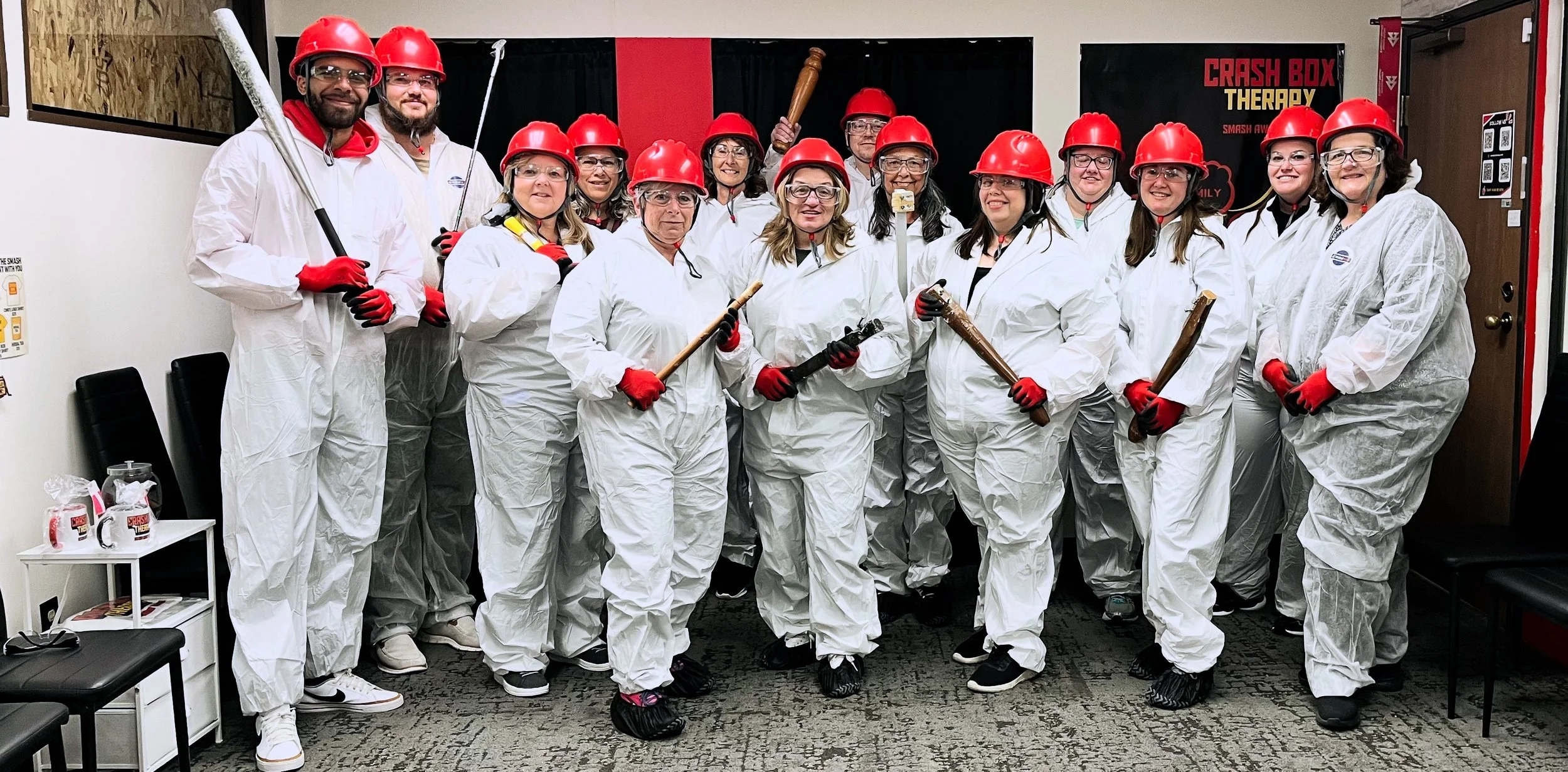 Group of people in a room dressed in white coveralls, red helmets, and gloves, holding various toy weapons, posing for a photo during a themed team activity.