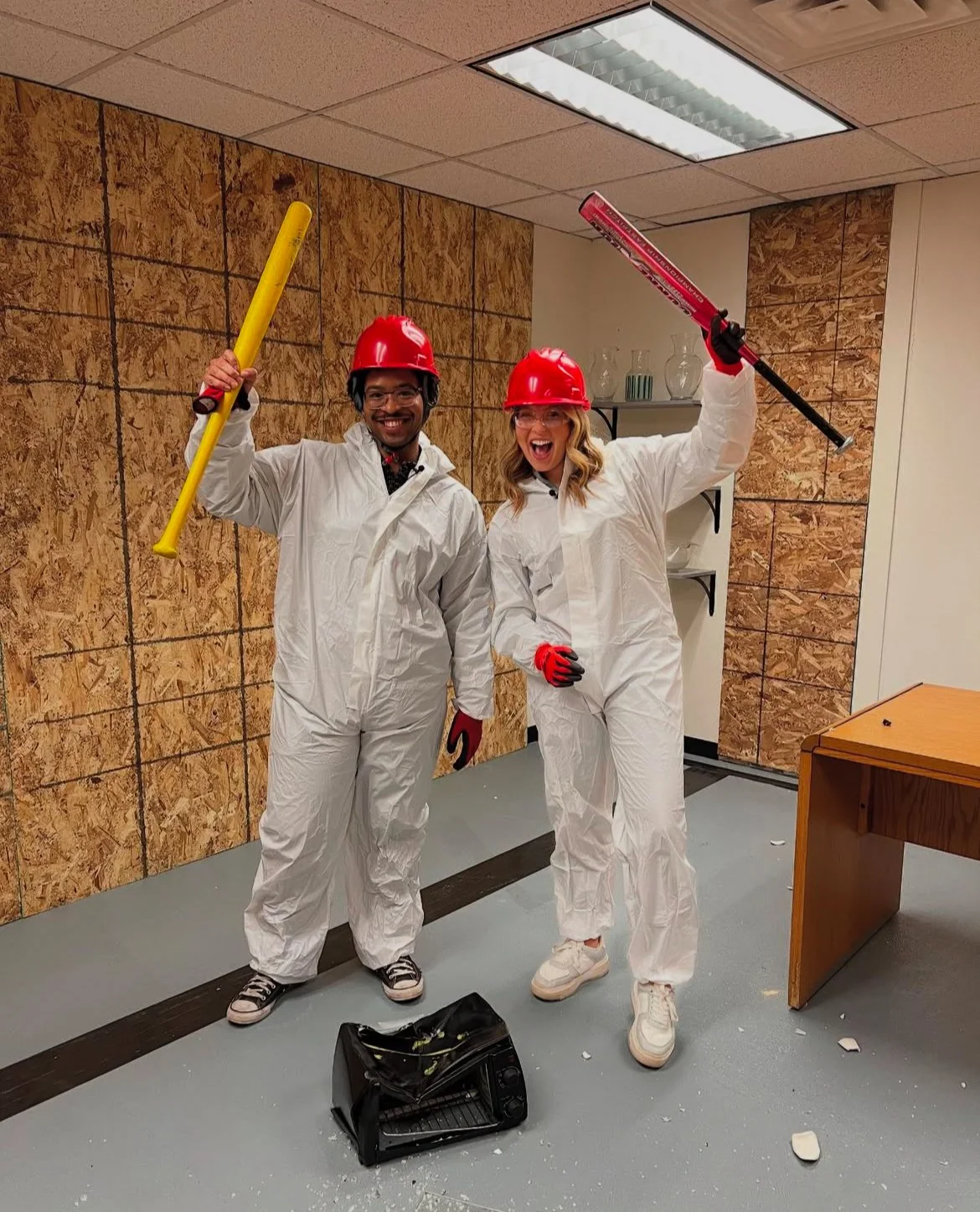 Two people posing in a room wearing white protective suits, red hard hats, and gloves. They are holding colorful baseball bats and smiling. A smashed object is on the floor, suggesting they are in a rage room.