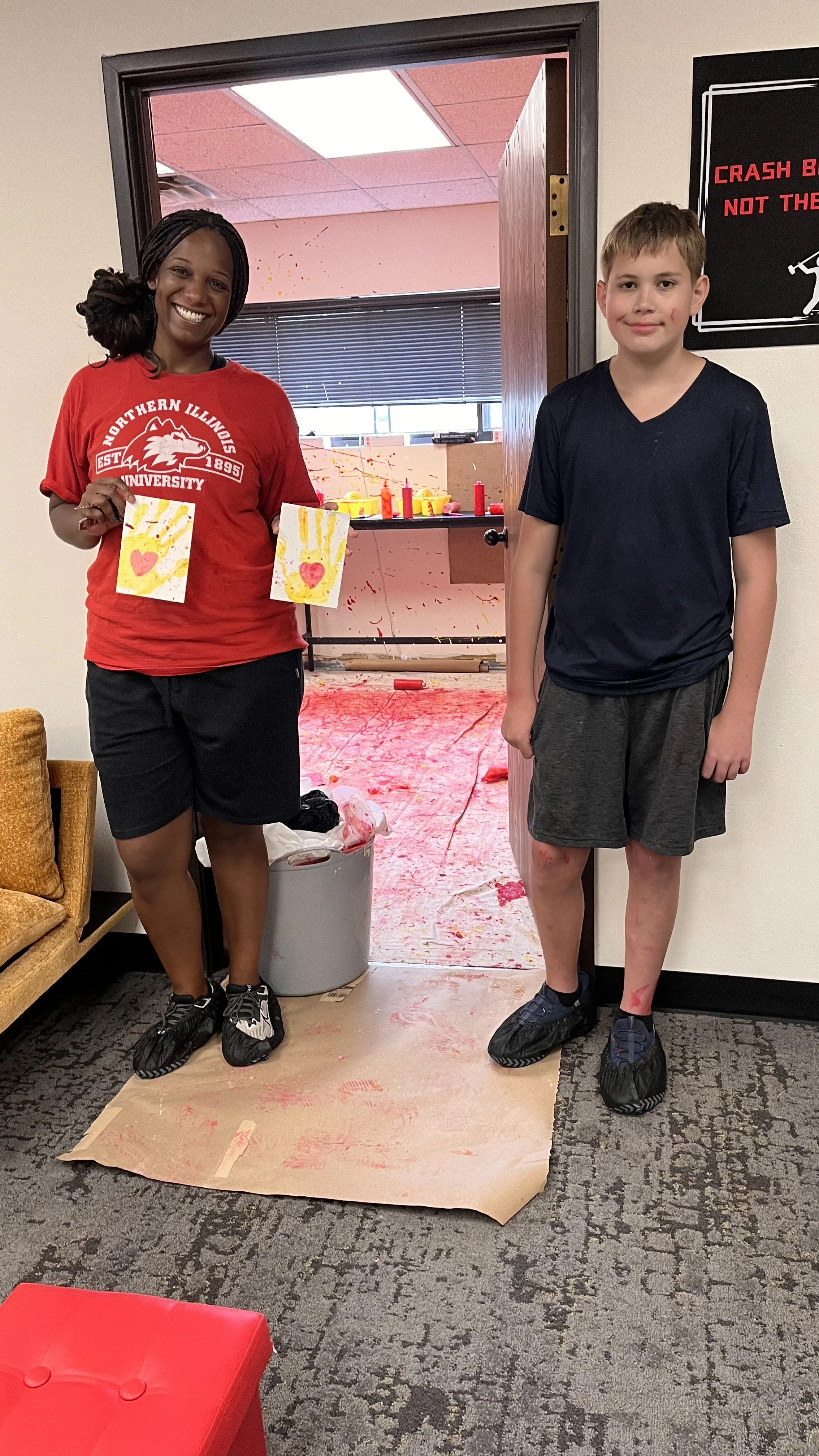 A woman and a boy standing in front of a messy room after painting. The woman is holding two pieces of artwork, smiling, while the boy stands beside her with a neutral expression. The room behind them has red paint splatters on the wall, floor, and c
