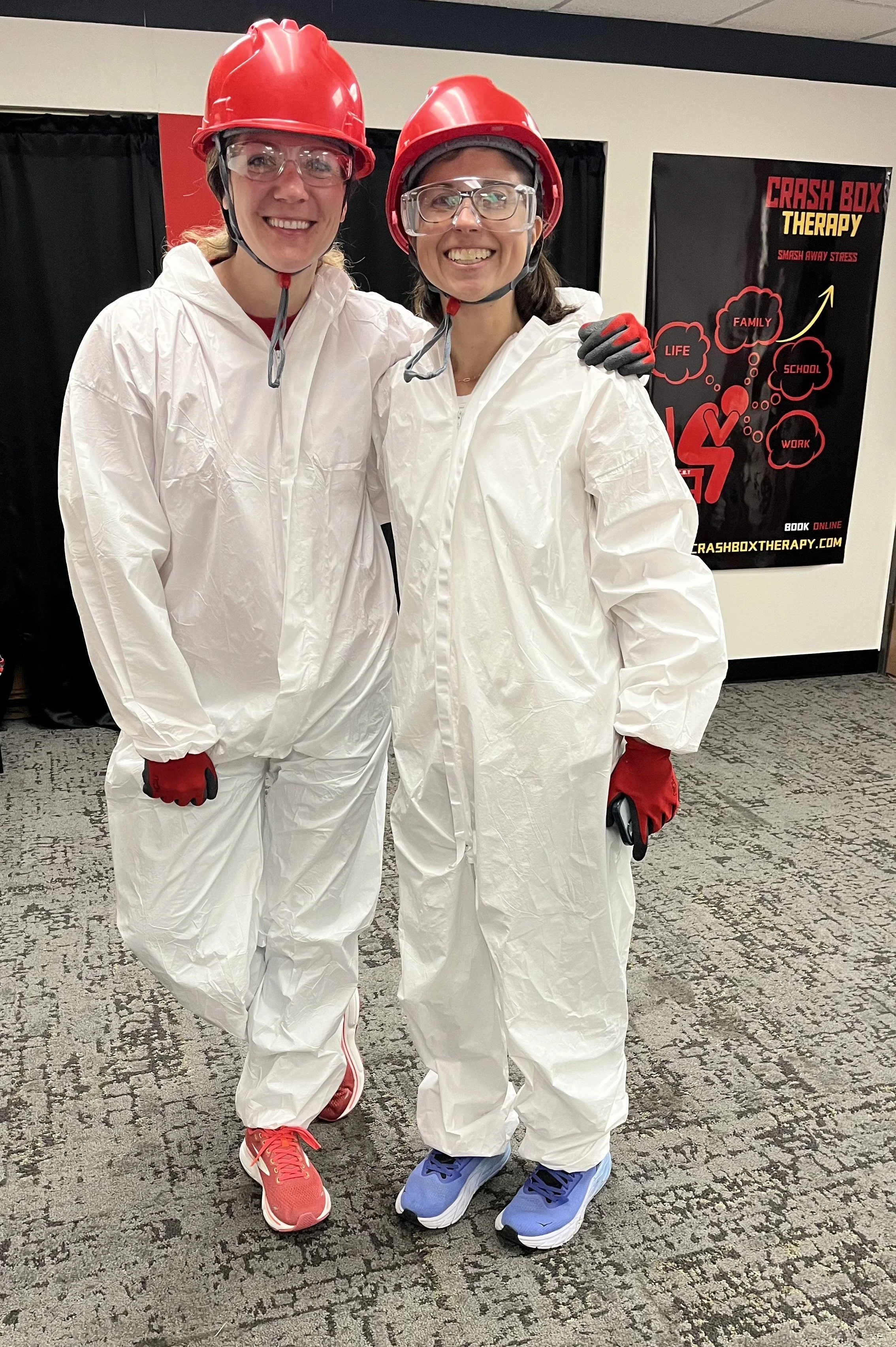 Two women wearing white jumpsuits, red helmets, and safety glasses, standing together and smiling, in an indoor setting with a poster behind them that reads 'Crash Box Therapy' and features a diagram with words like 'Life,' 'Family,' 'School,' 'Work,
