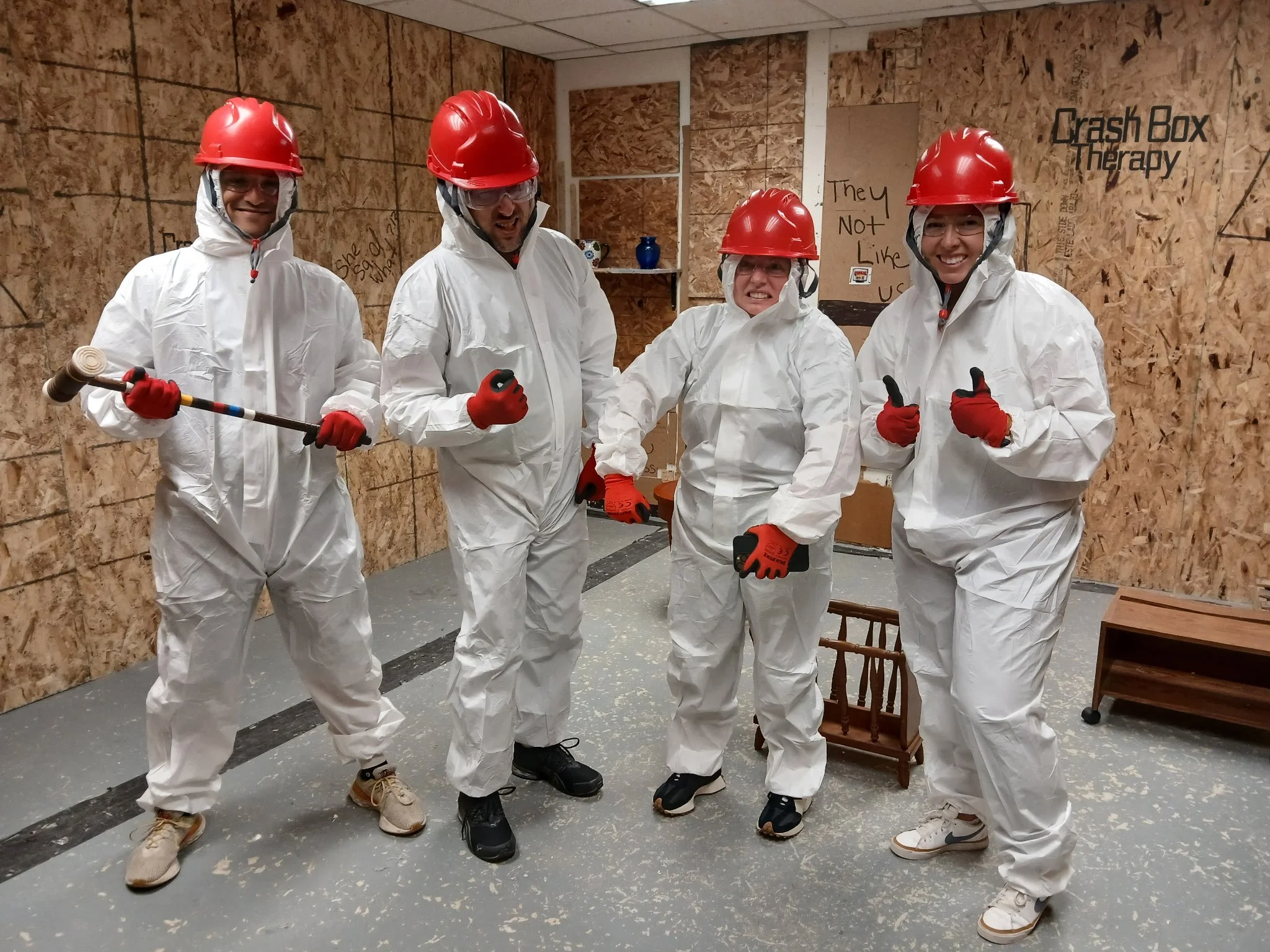 Four people dressed in white hazmat suits, red helmets, and gloves, standing inside a plywood-walled room, giving thumbs-up and grinning. One person holds a sledgehammer. The room has a sign that reads 'Crash Box Therapy' and a sign that says 'They N