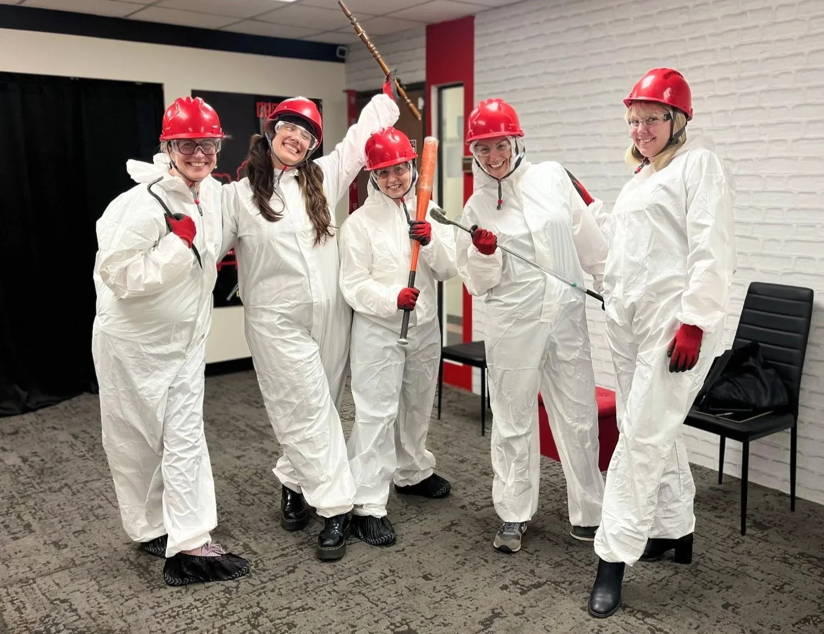 Five women in white protective suits, red helmets, and red gloves are smiling and holding tools, standing indoors against a white brick wall at Crash Box Therapy. 