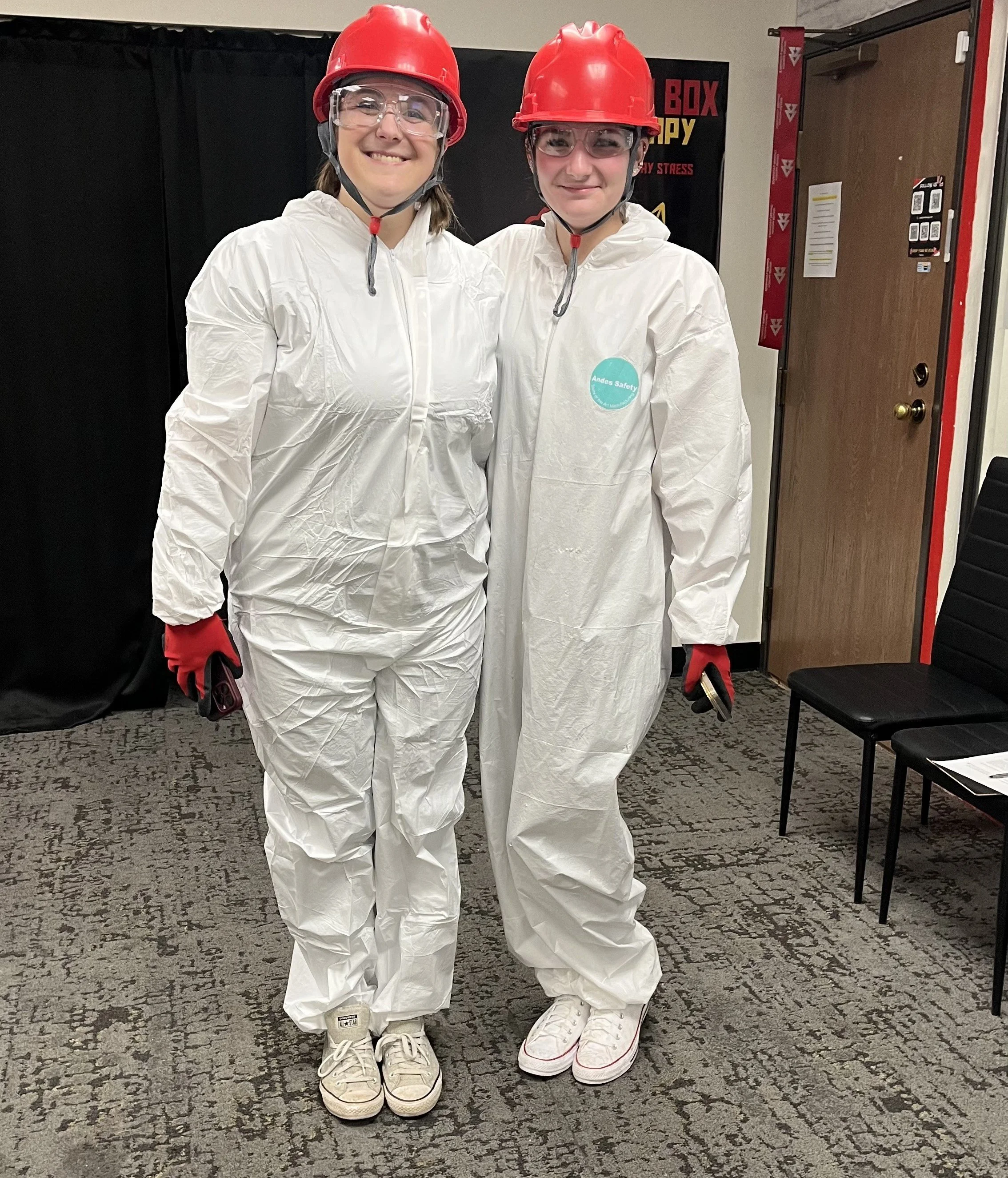 Two women in white protective suits, red helmets, and safety goggles, standing indoors on a gray patterned carpet, smiling at the camera.