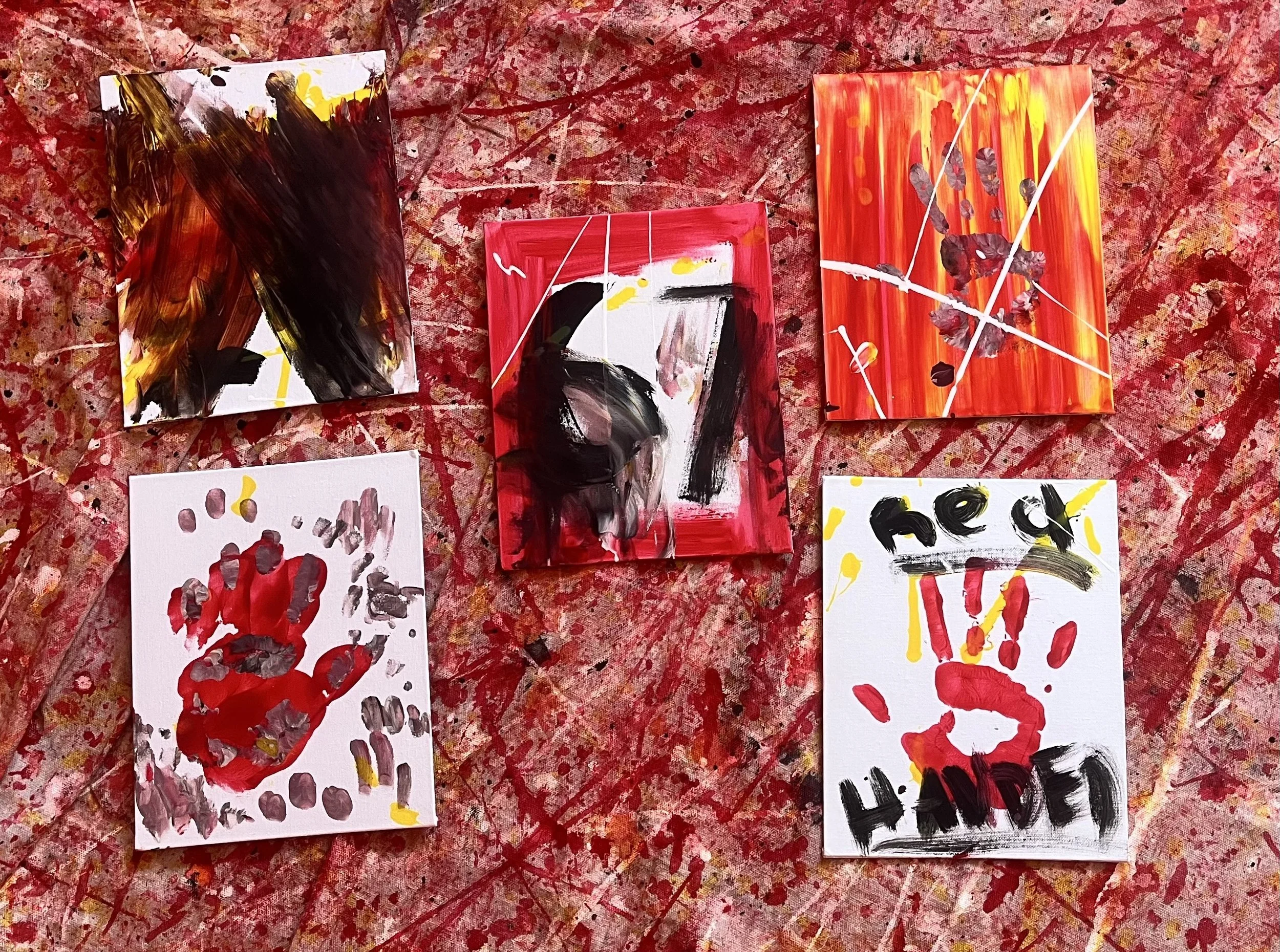 Five abstract paintings displayed on a splattered red and pink background. The paintings feature bold, expressive brushstrokes in black, red, yellow, and white, with some incorporating handprints and text.