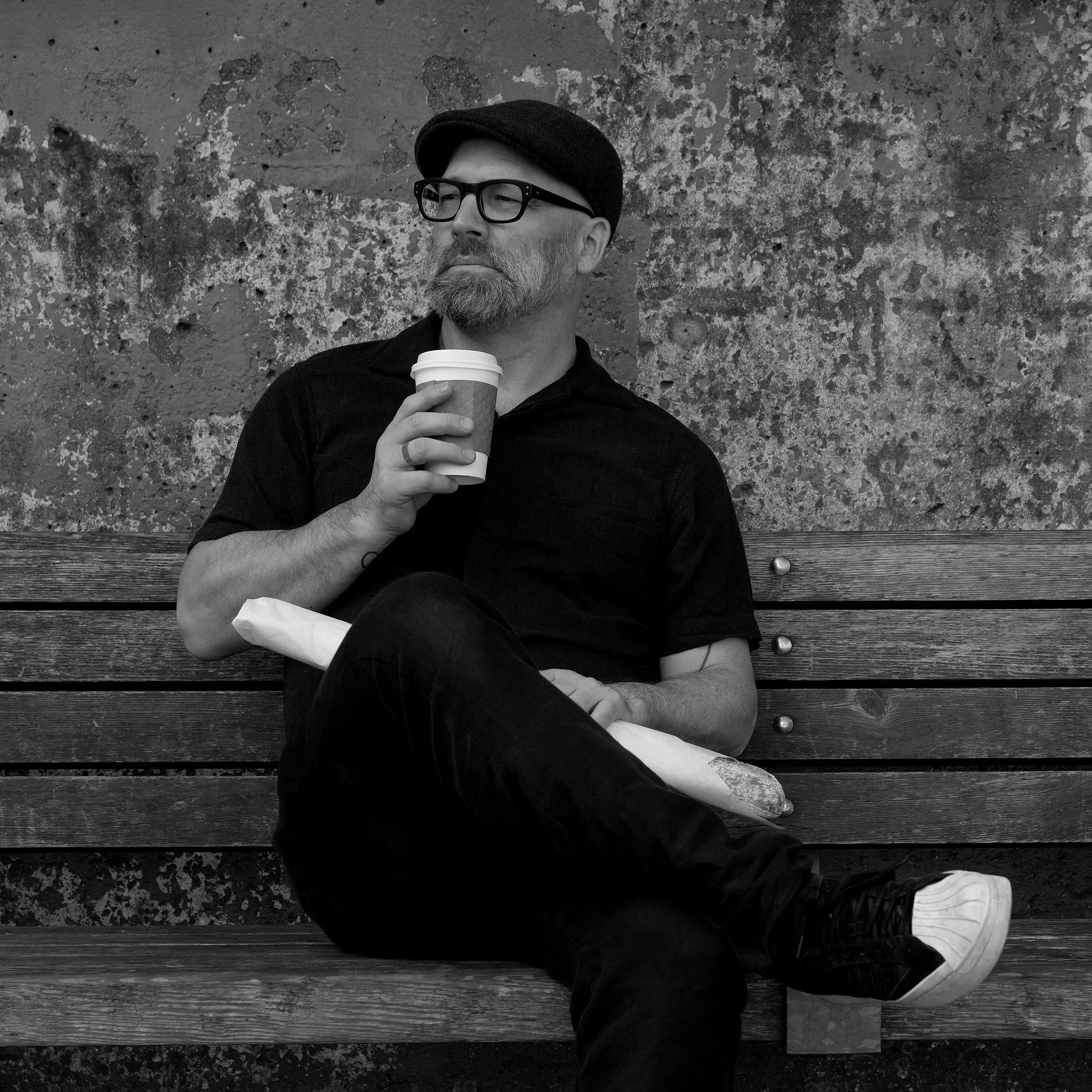 A man with a beard, glasses, wearing a hat, and dark clothing, sitting on a wooden bench against a textured wall. He is holding a coffee cup in one hand and a wrapped sandwich on his lap, with one leg crossed over the other.