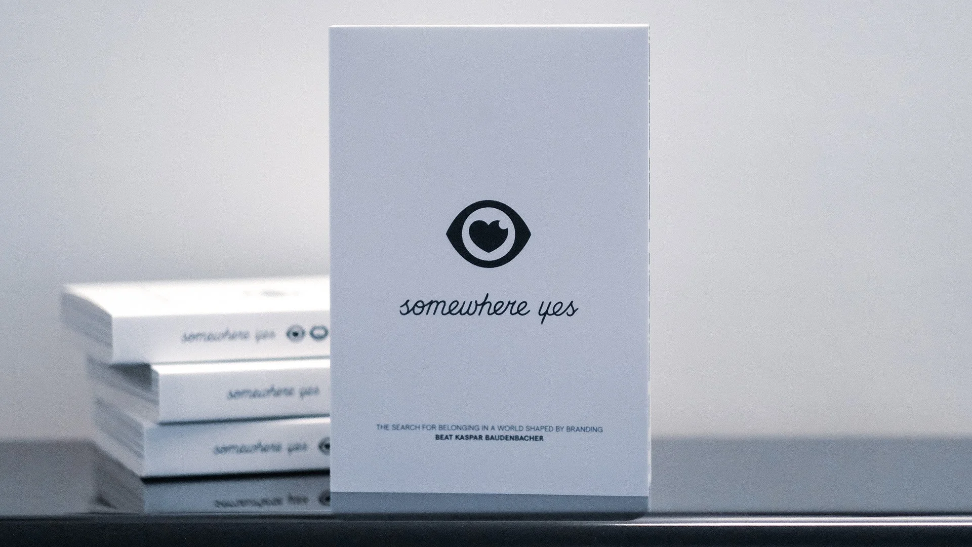 White book with black eye and heart logo, titled 'somewhere yes' and subtitle 'The search for belonging in a world shaped by branding' by Beat Kaspar Baudenbacher, standing upright on a reflective surface. Several similar books stacked behind it.