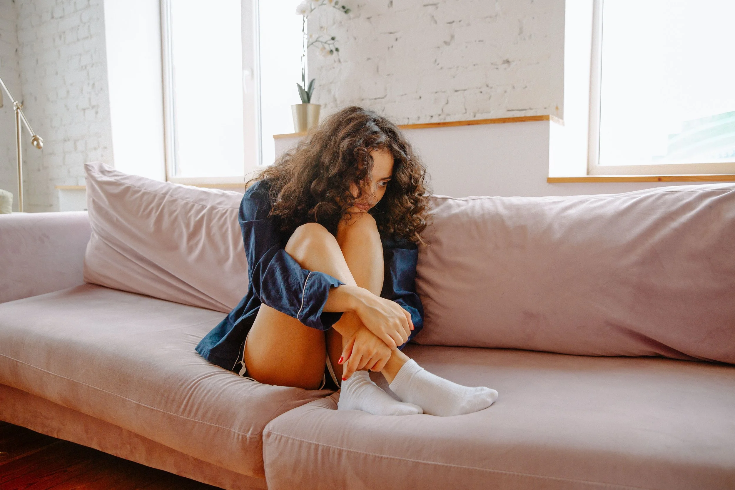 Woman with curly brown hair sitting curled up on a light pink couch, hugging her knees and looking downward in a bright living room.