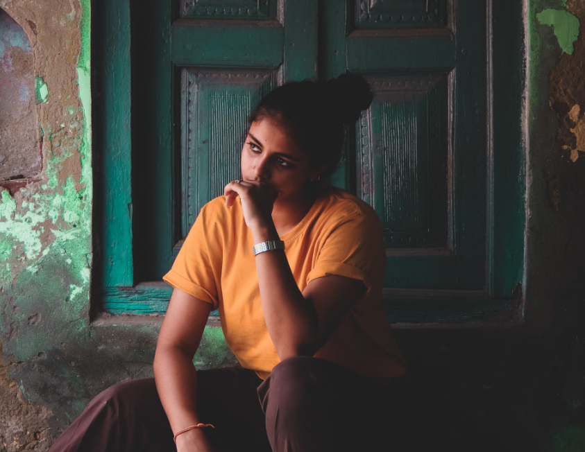 Woman wearing a mustard-yellow shirt sitting thoughtfully in front of a dark teal wooden door, resting her chin against her hand.