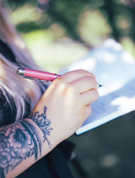 Close-up of a person with floral tattoos on their arm writing in a notebook with a pink pen outdoors.