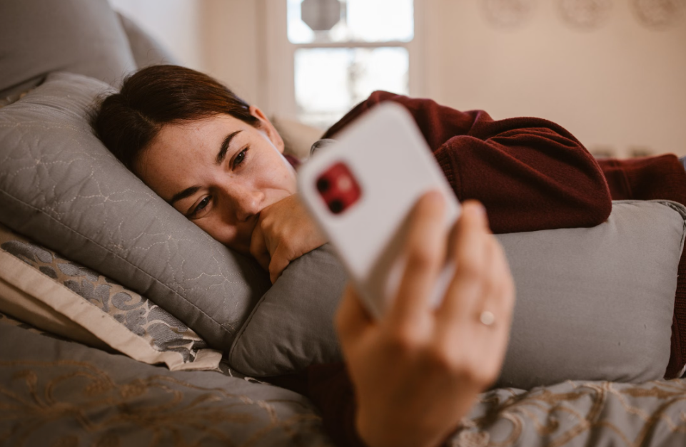 Woman lying in bed with her head resting on a pillow, looking at a smartphone while holding her hand near her mouth.