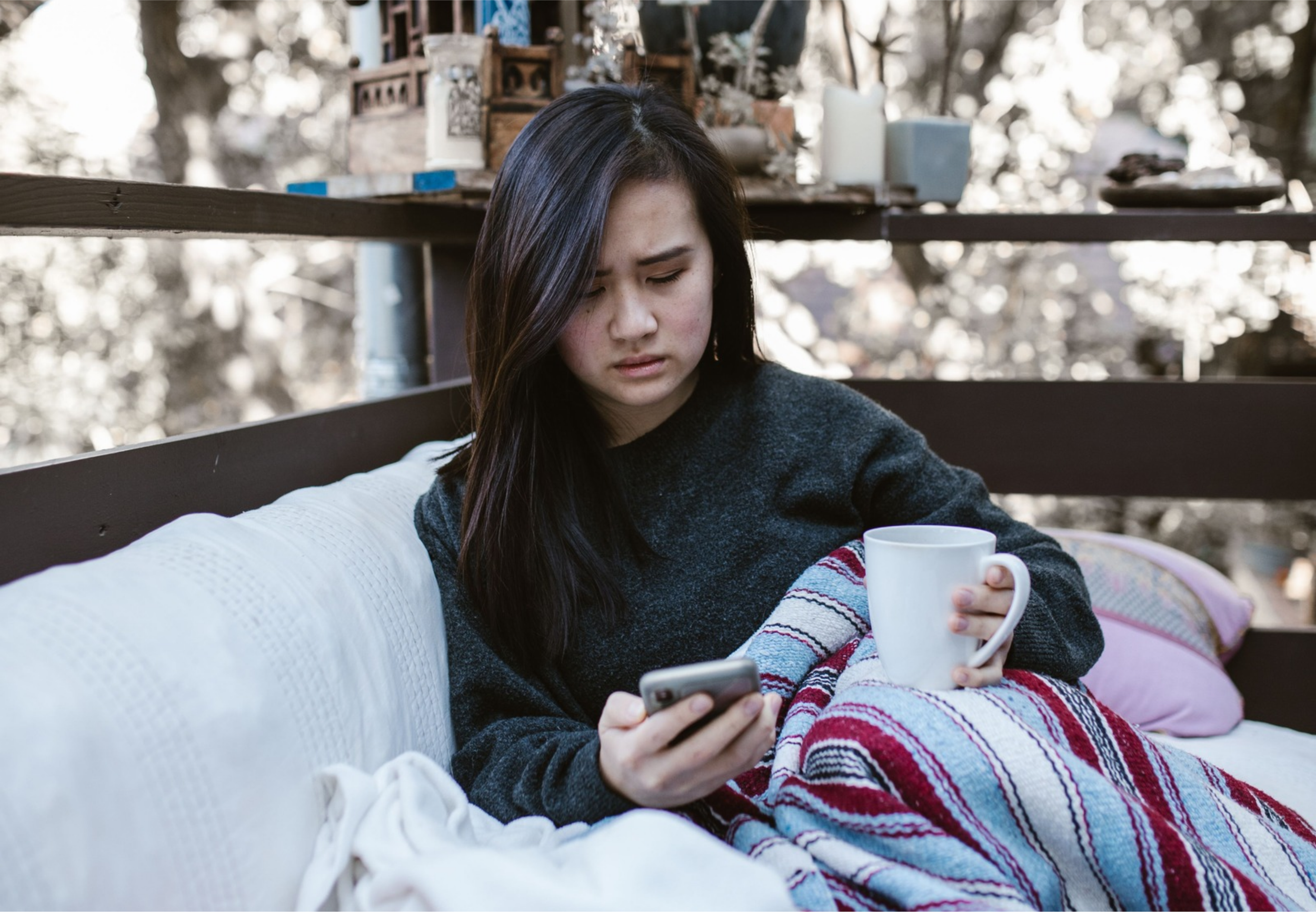 Woman sitting on a couch with a blanket and mug, looking thoughtfully at her phone.