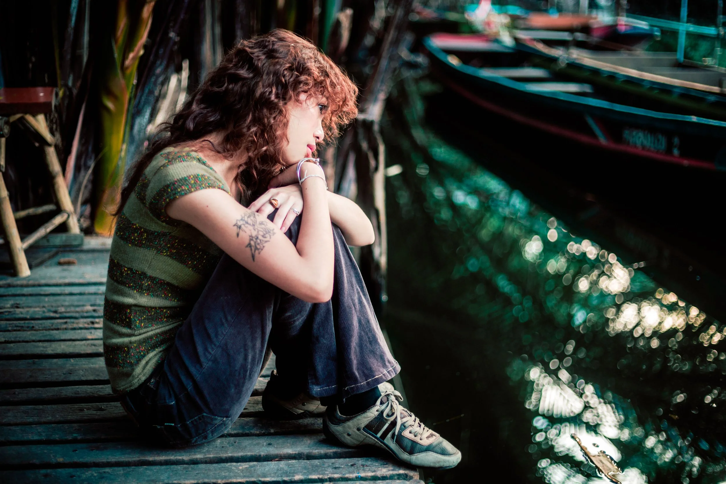 Woman with curly brown hair and an arm tattoo sitting on a wooden dock beside the water, resting her chin on her folded arms and looking thoughtfully into the distance.