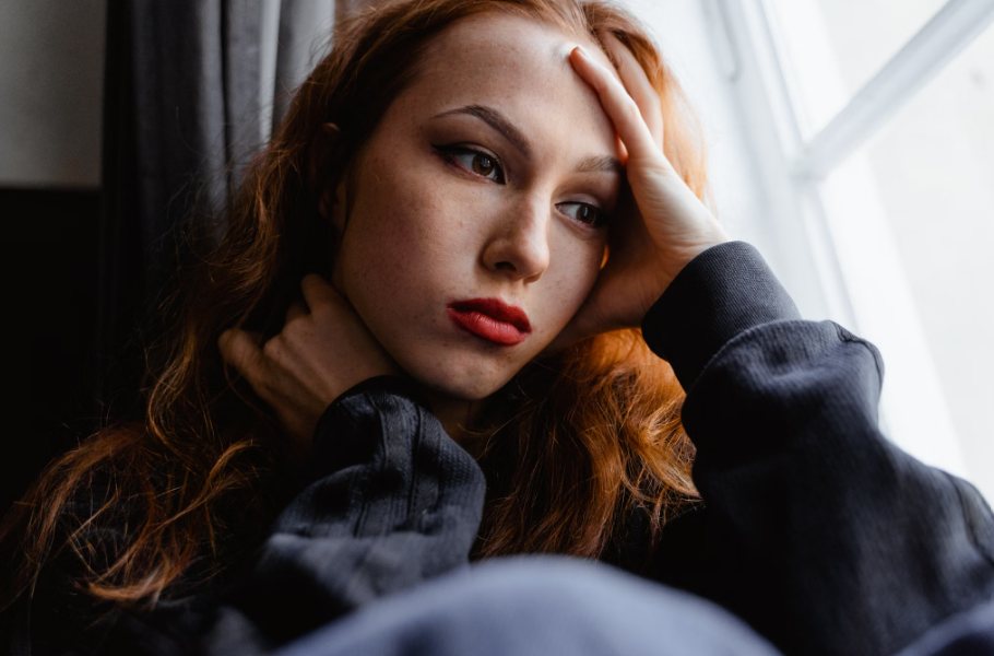 Red-haired woman sitting by a window with her hand resting against her forehead, looking thoughtful and emotionally tired.