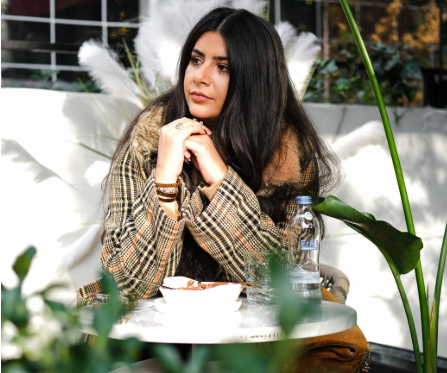 Woman with long dark hair wearing a plaid jacket, sitting at an outdoor café table with her hands beneath her chin and looking thoughtfully into the distance.
