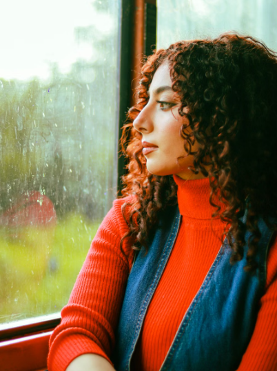 Woman with curly hair wearing an orange turtleneck, gazing thoughtfully out a rain-speckled window.