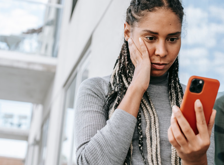 Woman with braided hair holding an orange smartphone near a window, looking concerned and thoughtful.