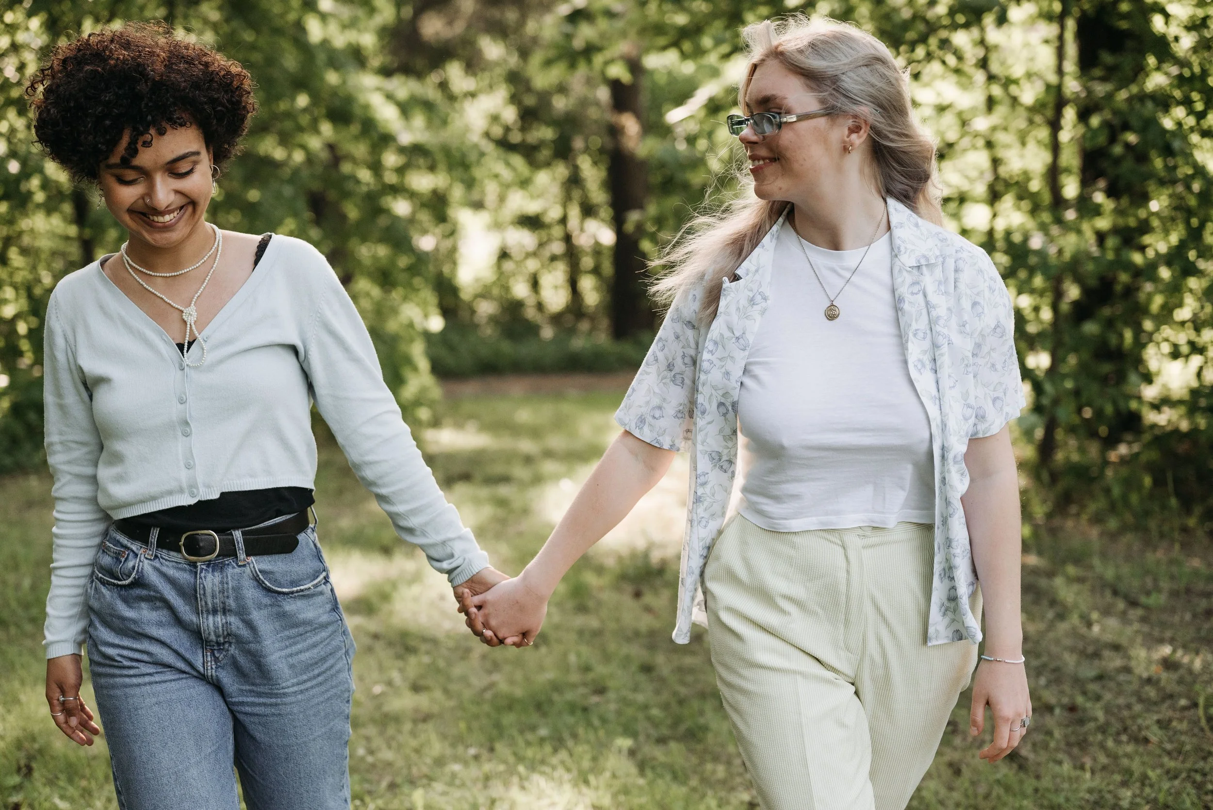 Two women smiling and holding hands while walking together through a green, wooded area.