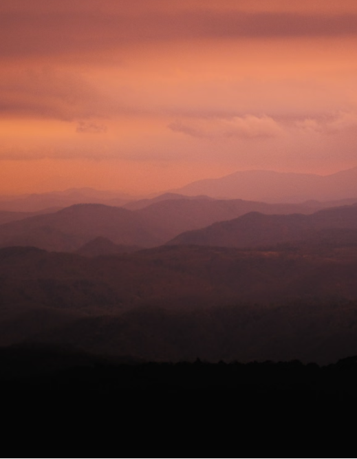 Warm sunset over layered mountain ridges in soft pink and plum tones.