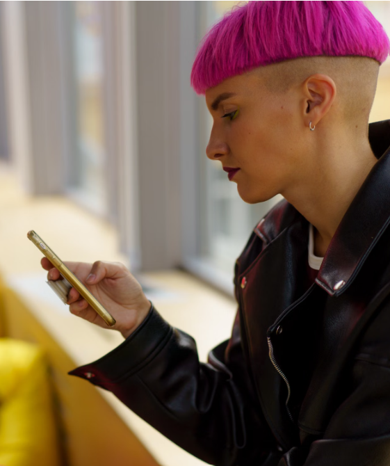 Person with bright pink hair and shaved sides wearing a black leather jacket, looking down at a smartphone.