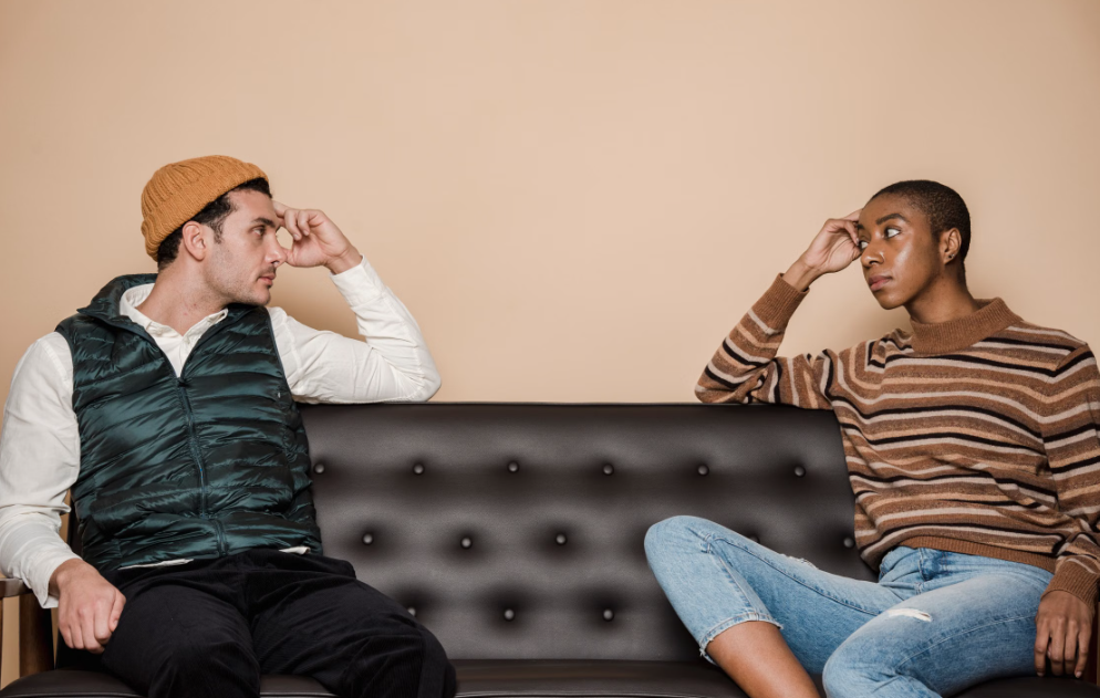 Two people sitting at opposite ends of a black couch, looking at each other with serious expressions against a beige background.