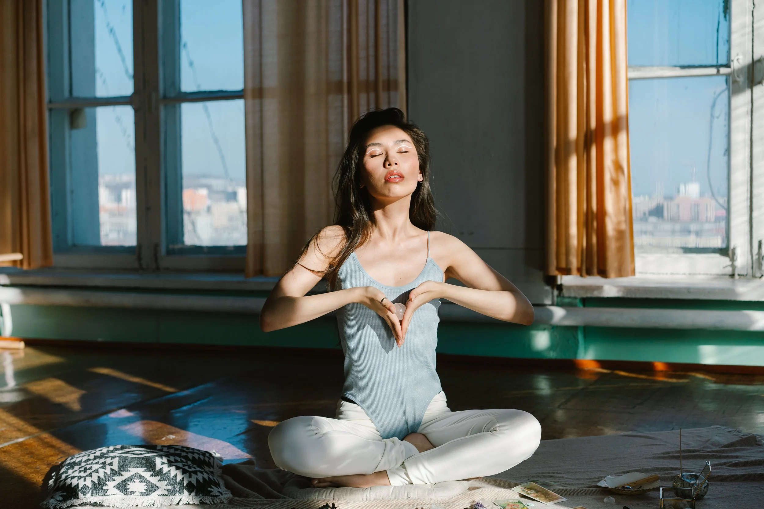 Woman with long dark hair sitting cross-legged on a mat in a sunlit room, eyes closed and hands positioned together in front of her chest.