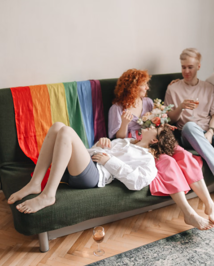 Three people relaxing together on a couch with a rainbow Pride flag draped over the back, surrounded by flowers in a cozy living room.