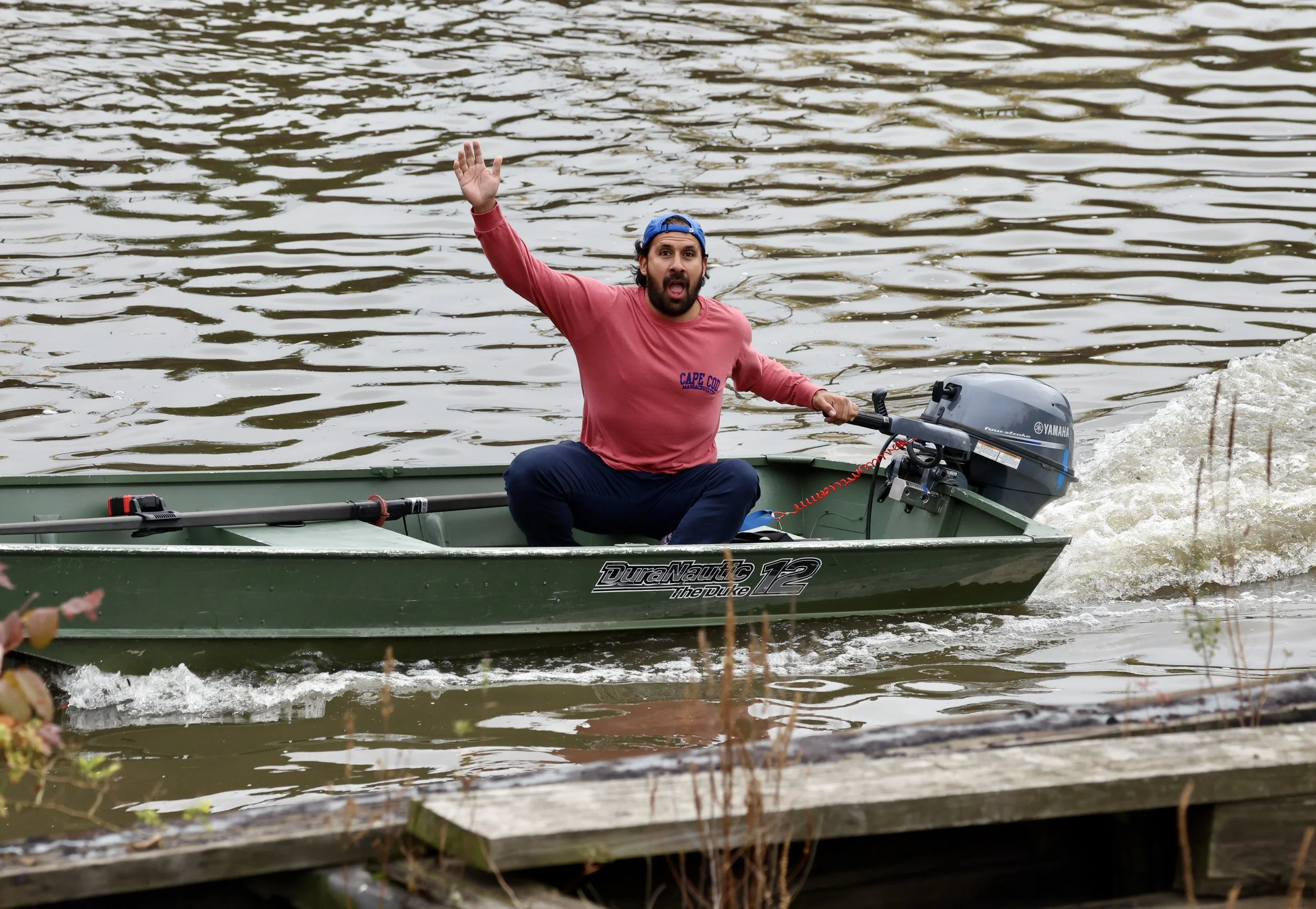 A man in a red shirt and blue cap sitting in a small green motor boat on a river, waving with his left hand, with a Yamaha outboard motor attached to the boat.