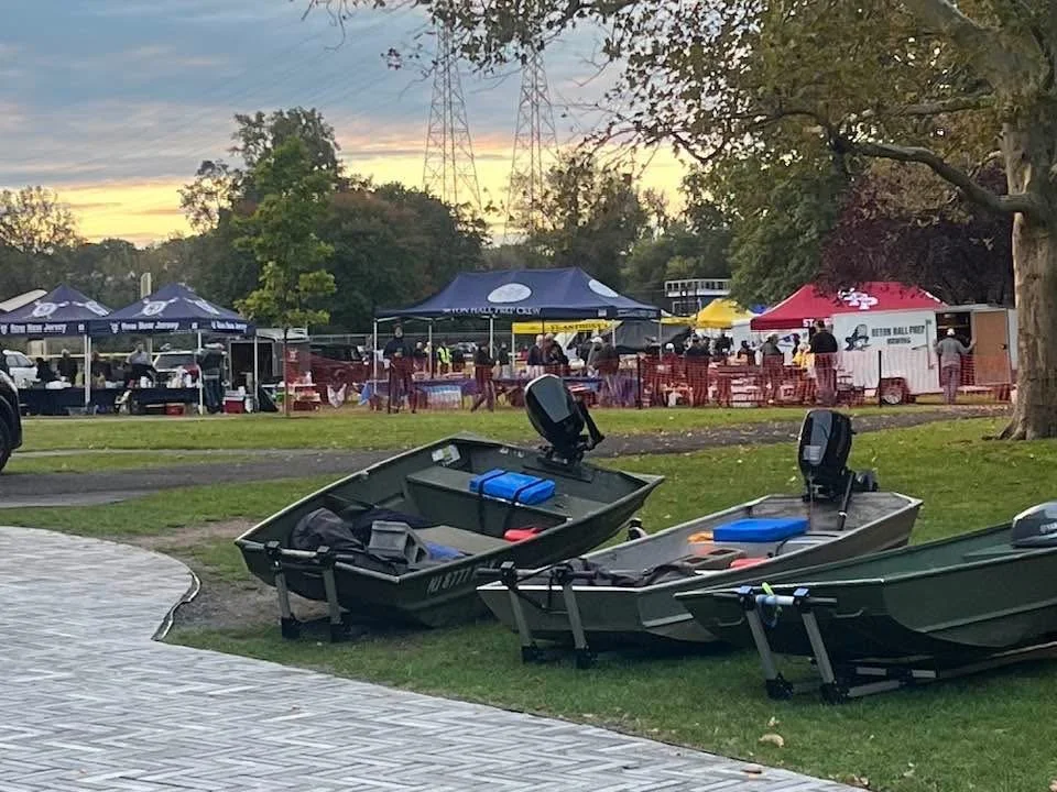 Three small boats parked on a grassy area near a paved sidewalk, with tents and people gathering in the background during sunset, surrounded by trees and electrical towers.