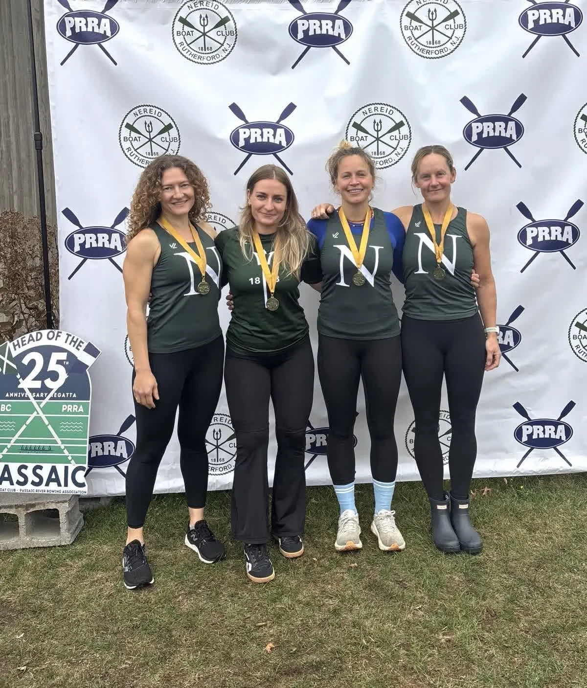 Four women standing side by side outdoors, smiling, wearing matching green athletic tank tops with white 'N' and black pants, medals around their necks, in front of a backdrop with rowing and rowing club logos.