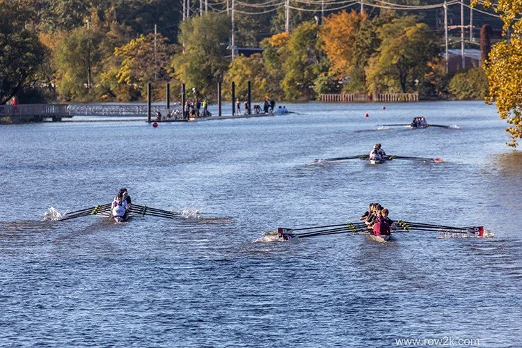 Rowers in a boat race on a river with trees displaying autumn colors in the background