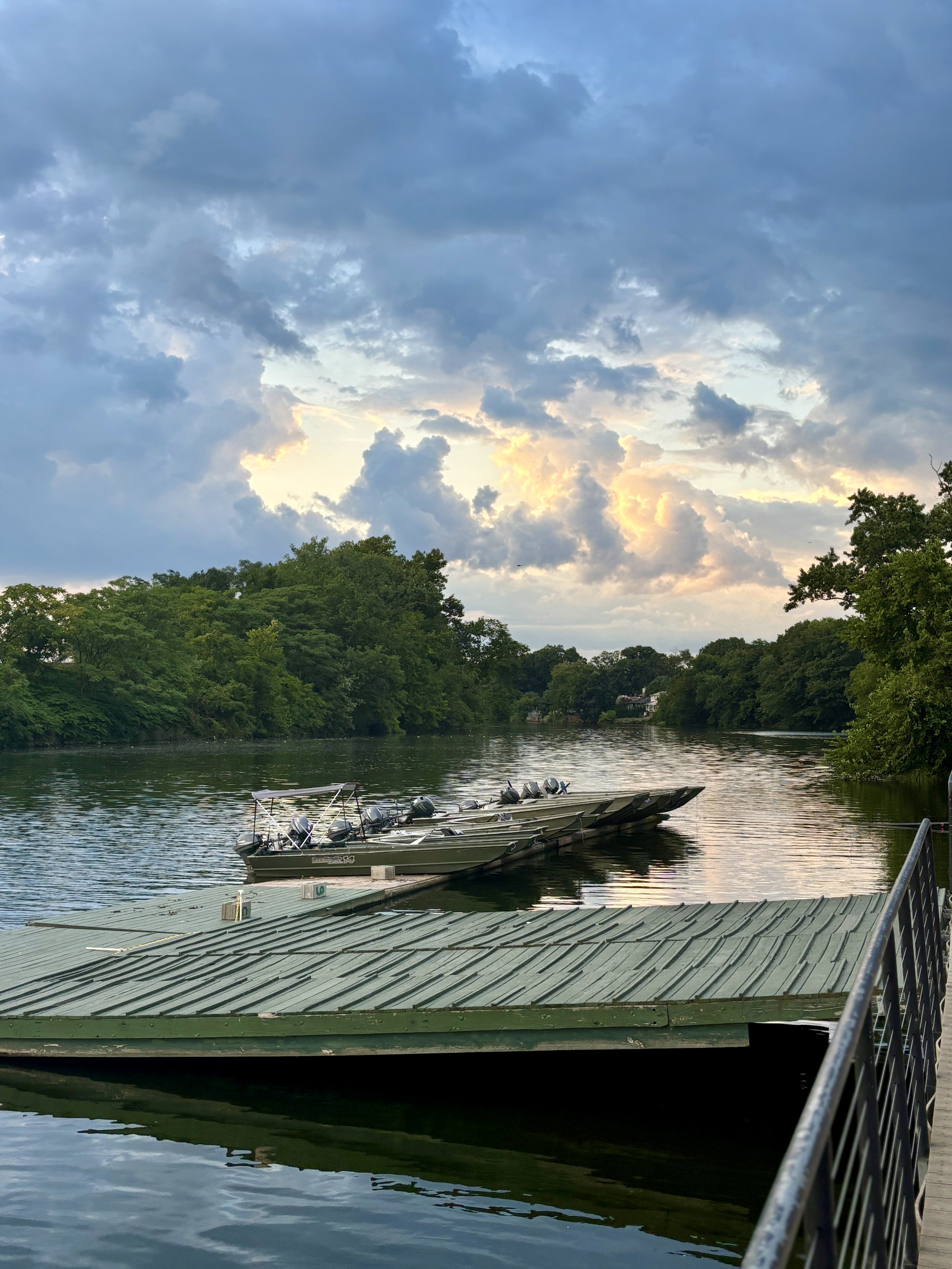 Boats docked on a calm river with a green floating dock in the foreground, lush trees lining the riverbank, and a partly cloudy sky with hints of sunlight at sunset.