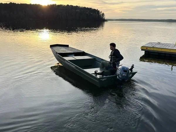 A man standing in a small motorboat on a lake at sunset with a floating dock nearby.