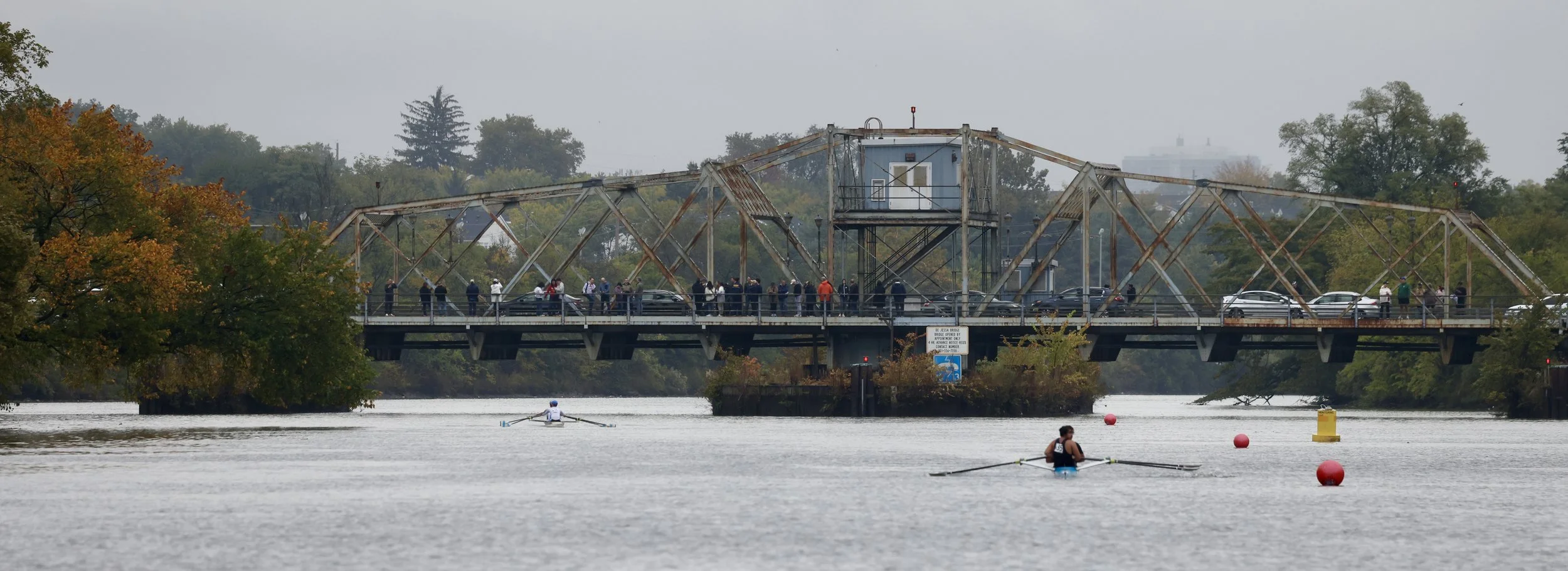 View of a river with two rowers in boats, a rusty steel bridge with pedestrian and vehicle traffic, and fall foliage on trees in the background.