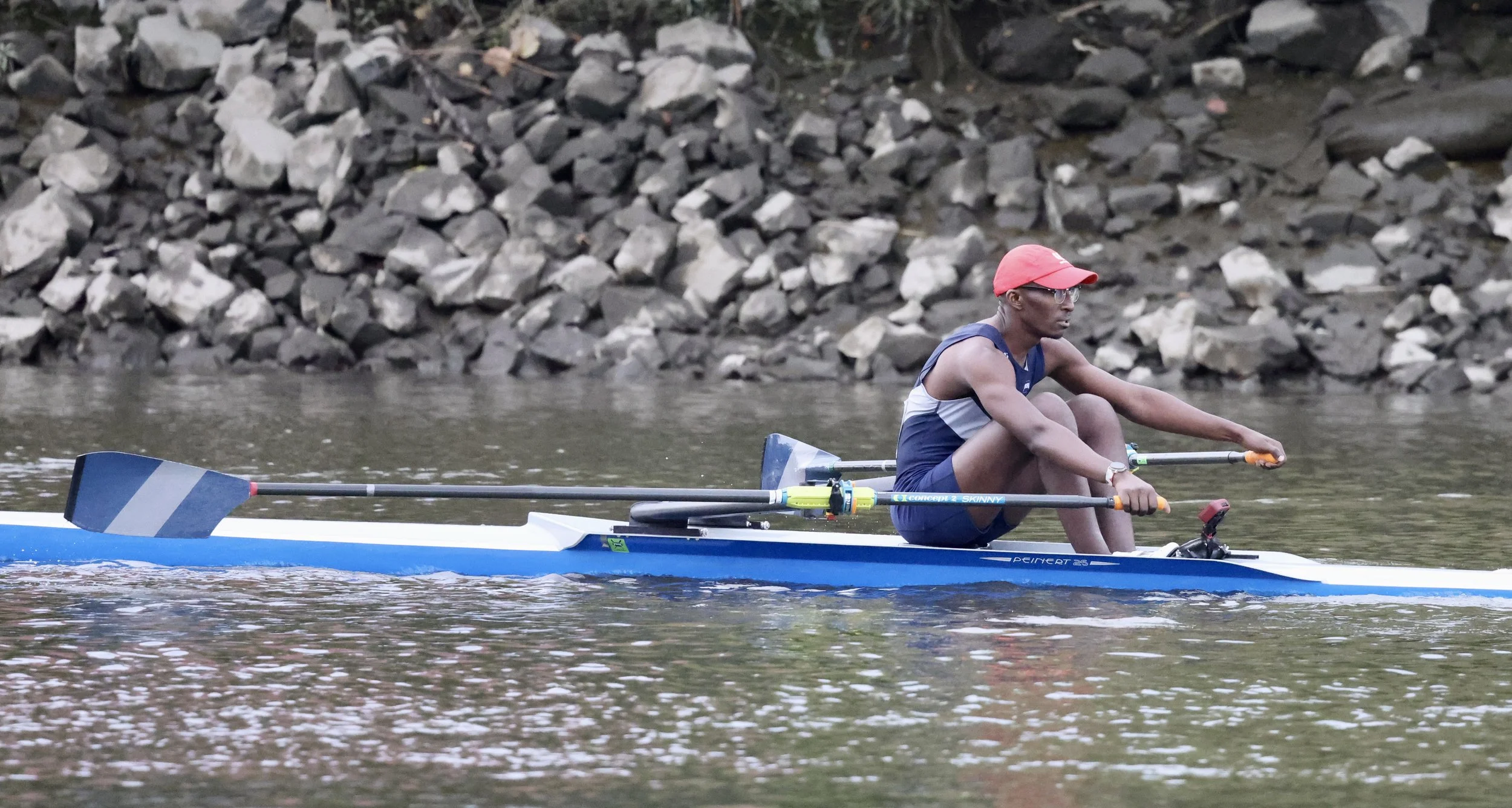 A person in athletic clothing, including a red cap, sitting in a boat on a river, holding oars, with a rocky riverbank in the background.