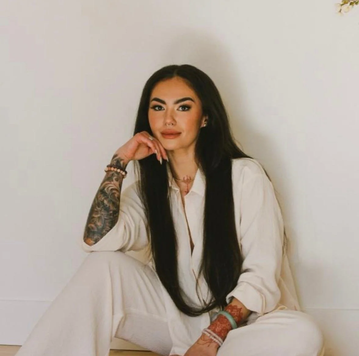 Woman with long black hair, tattoos, and jewelry posing in a white outfit against a plain wall