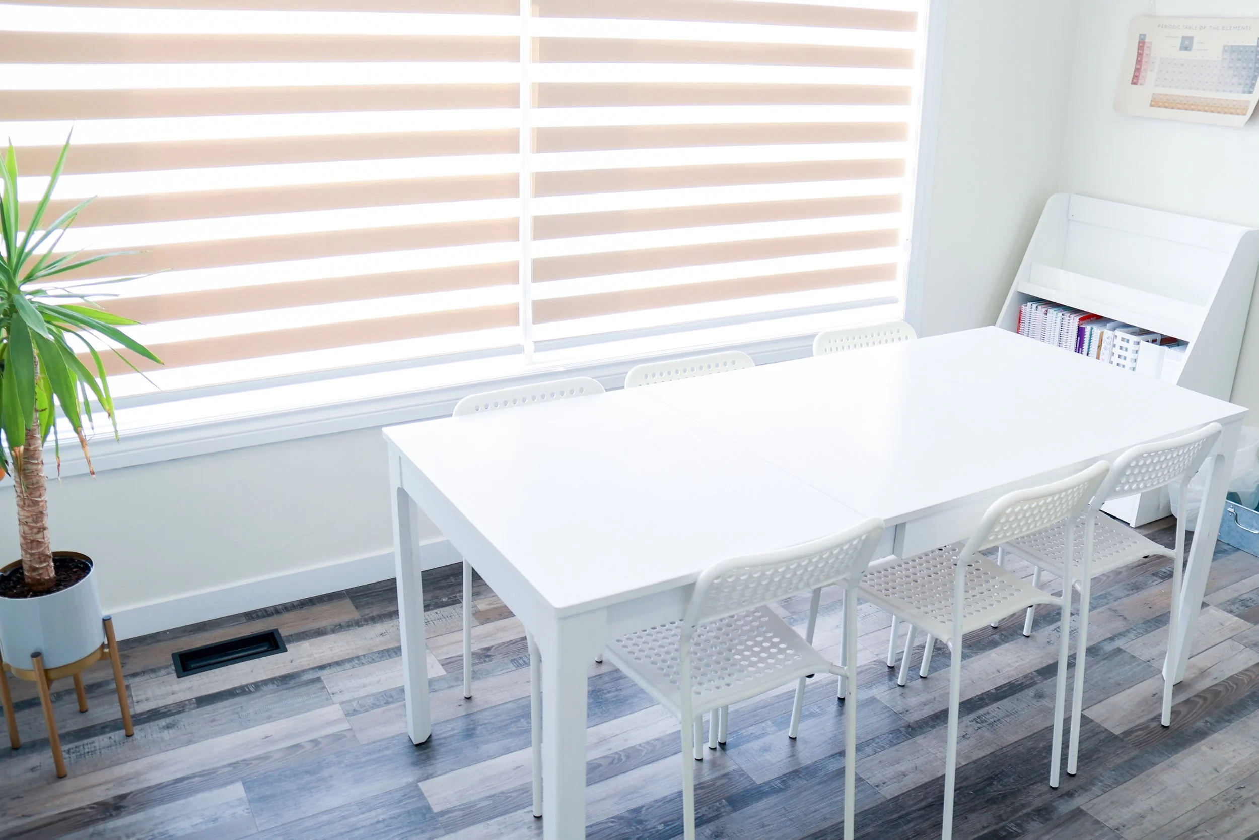 Bright meeting room with a white table, six white chairs, floor-to-ceiling window with beige blinds, bookshelf with books, potted plant in a gray pot, and a periodic table poster on the wall.