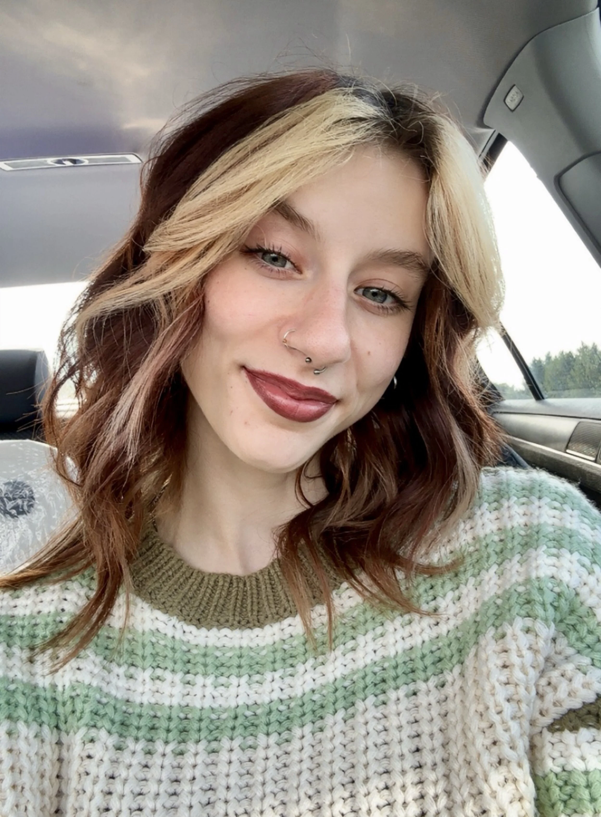 A young woman with glasses and piercings smiling in a cozy indoor setting.
