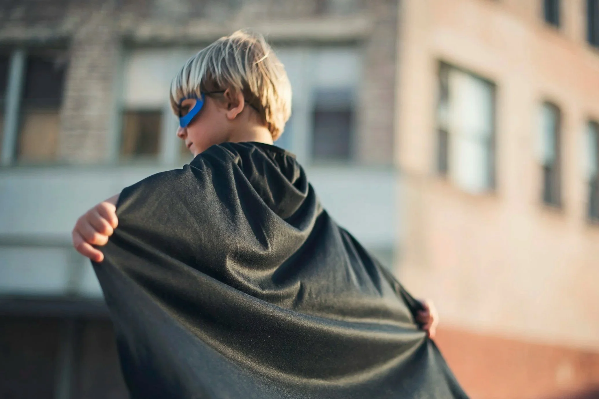 A young boy wearing a blue superhero mask and a blue cape, smiling widely, in a room with wooden shelves and books, celebrating a birthday event.